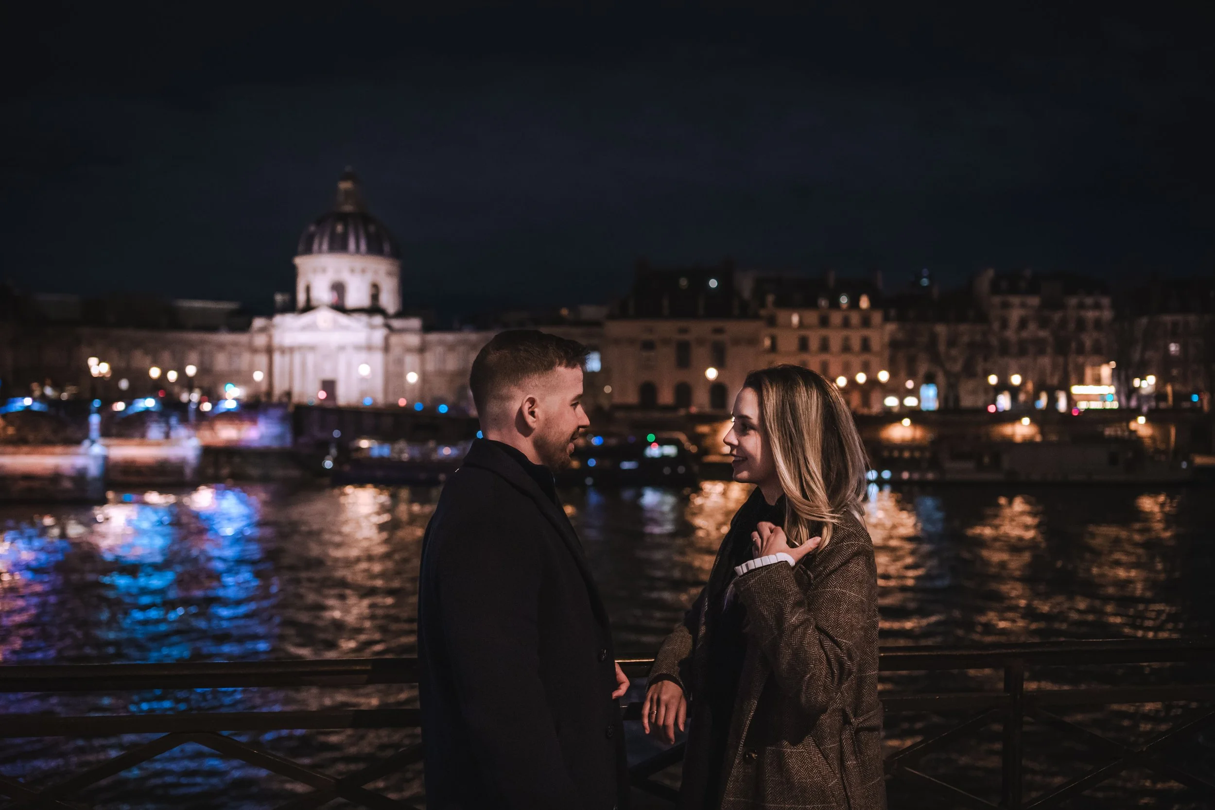 Un homme et une femme se regardent et sourient près de la Seine à Paris la nuit, avec le pont des Arts et des bâtiments illuminés en arrière-plan.
