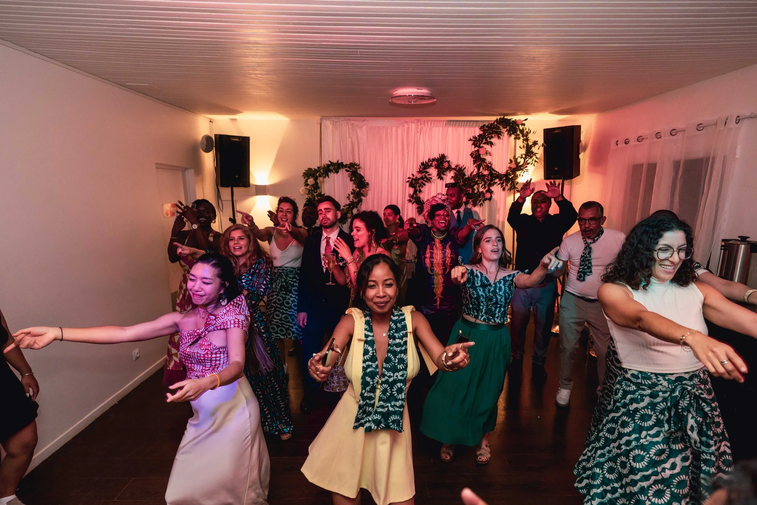 Groupe de personnes dansant et s'amusant lors d'une fête ou d'une célébration dans une salle avec décor floral et éclairage tamisé.
