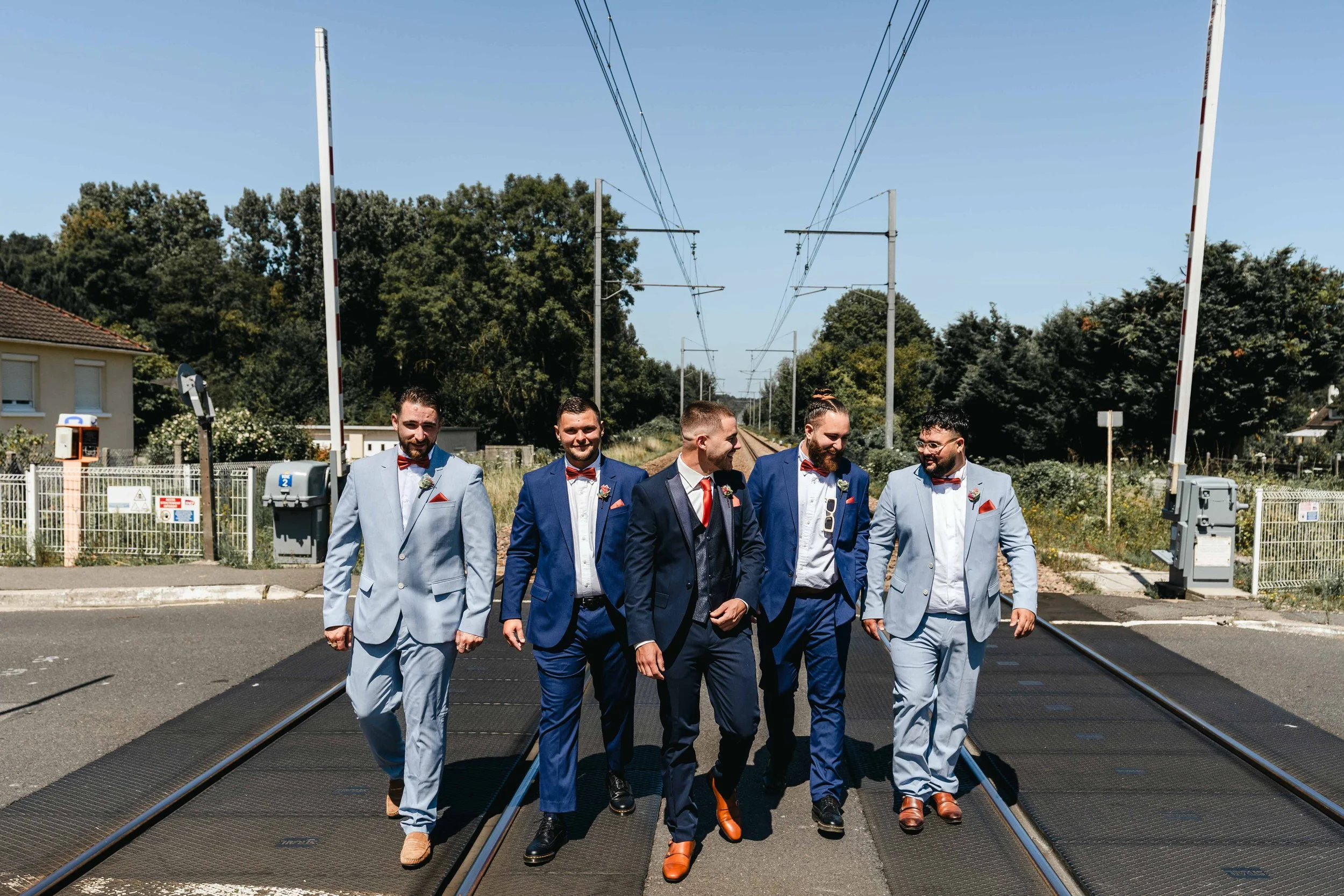 Groupe de cinq hommes habillés en costumes de mariage, marchant sur un passage à niveau sous un ciel clair.