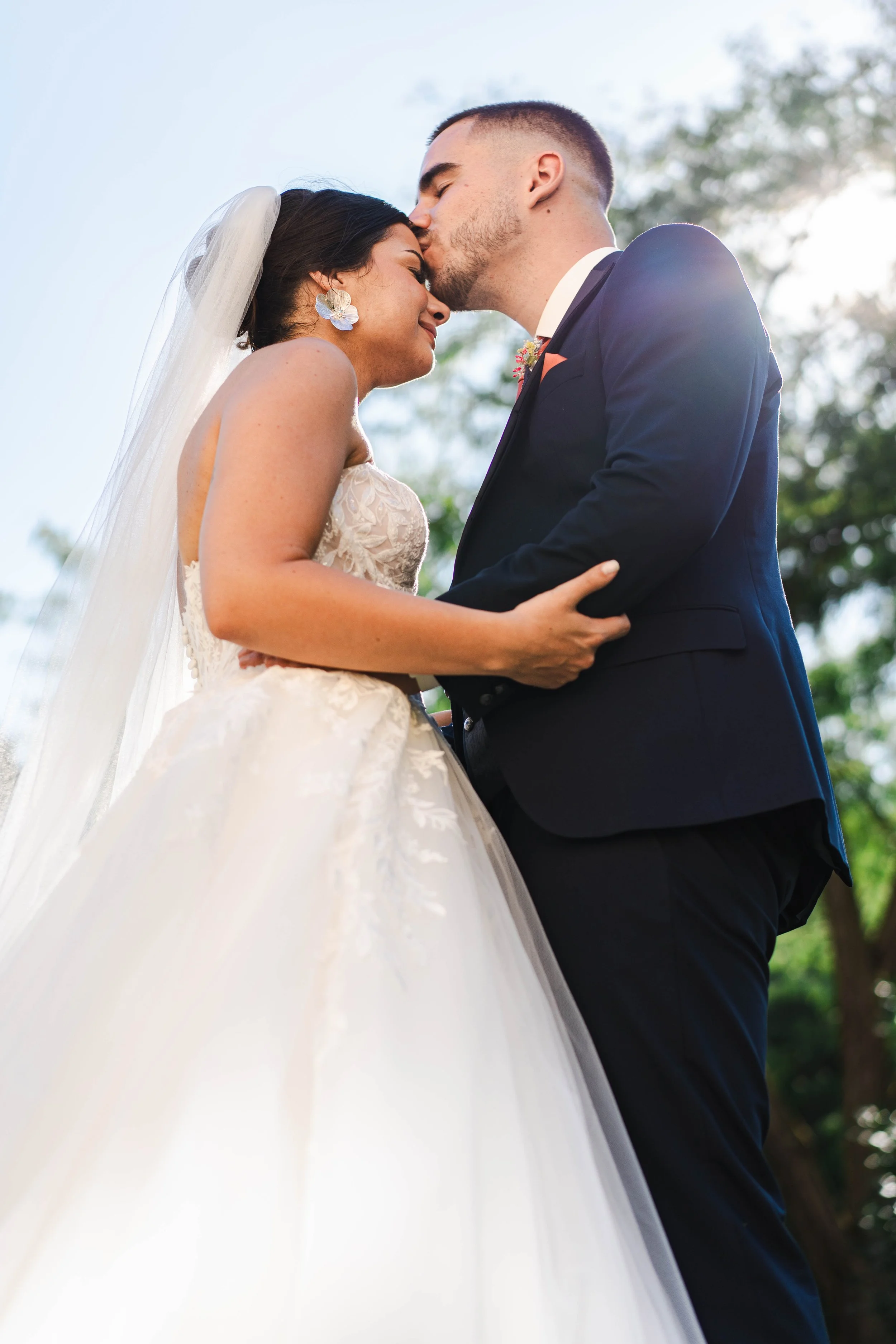 Un couple de mariés, un homme et une femme, s'embrassent tendrement en plein air, avec des arbres en arrière-plan.