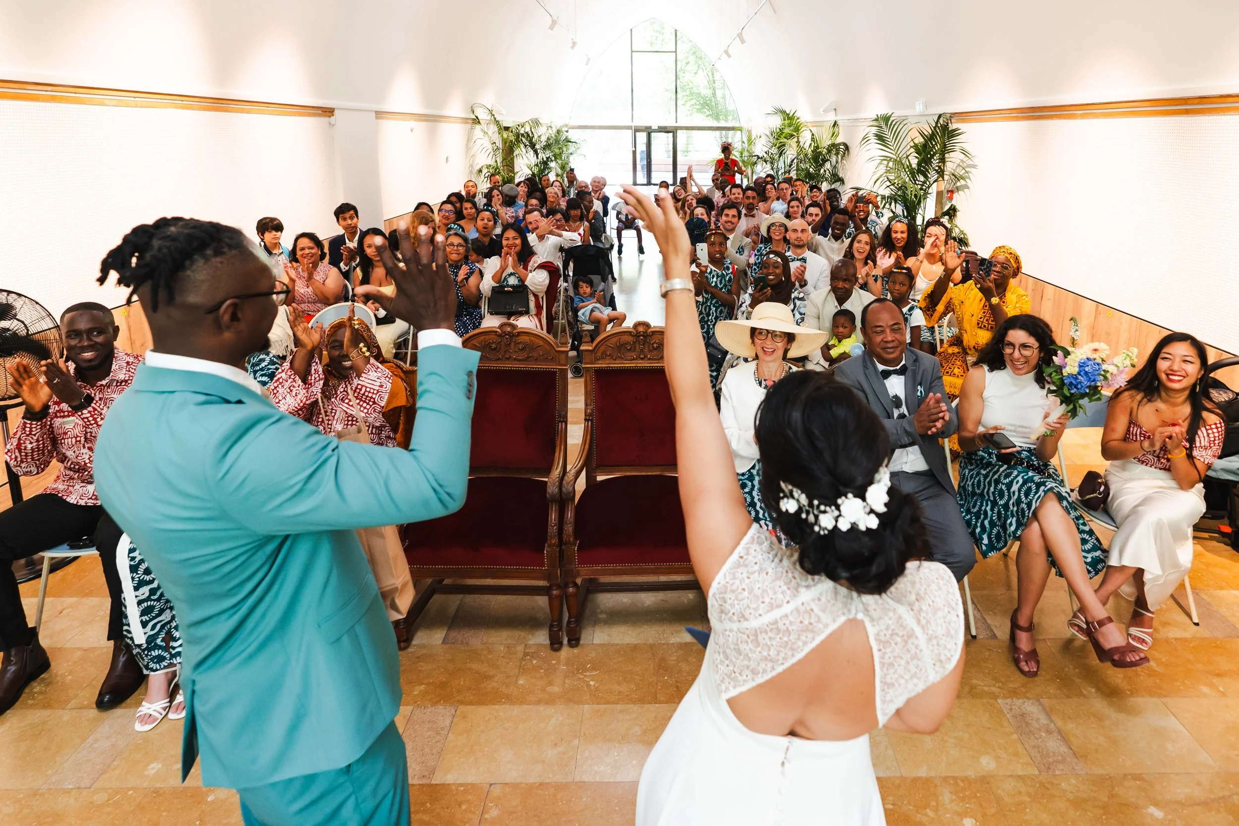 Un couple de mariés au centre de l'image, entouré d'invités lors d'une cérémonie de mariage, dans une salle bien éclairée avec des plantes vertes et des personnes souriantes.
