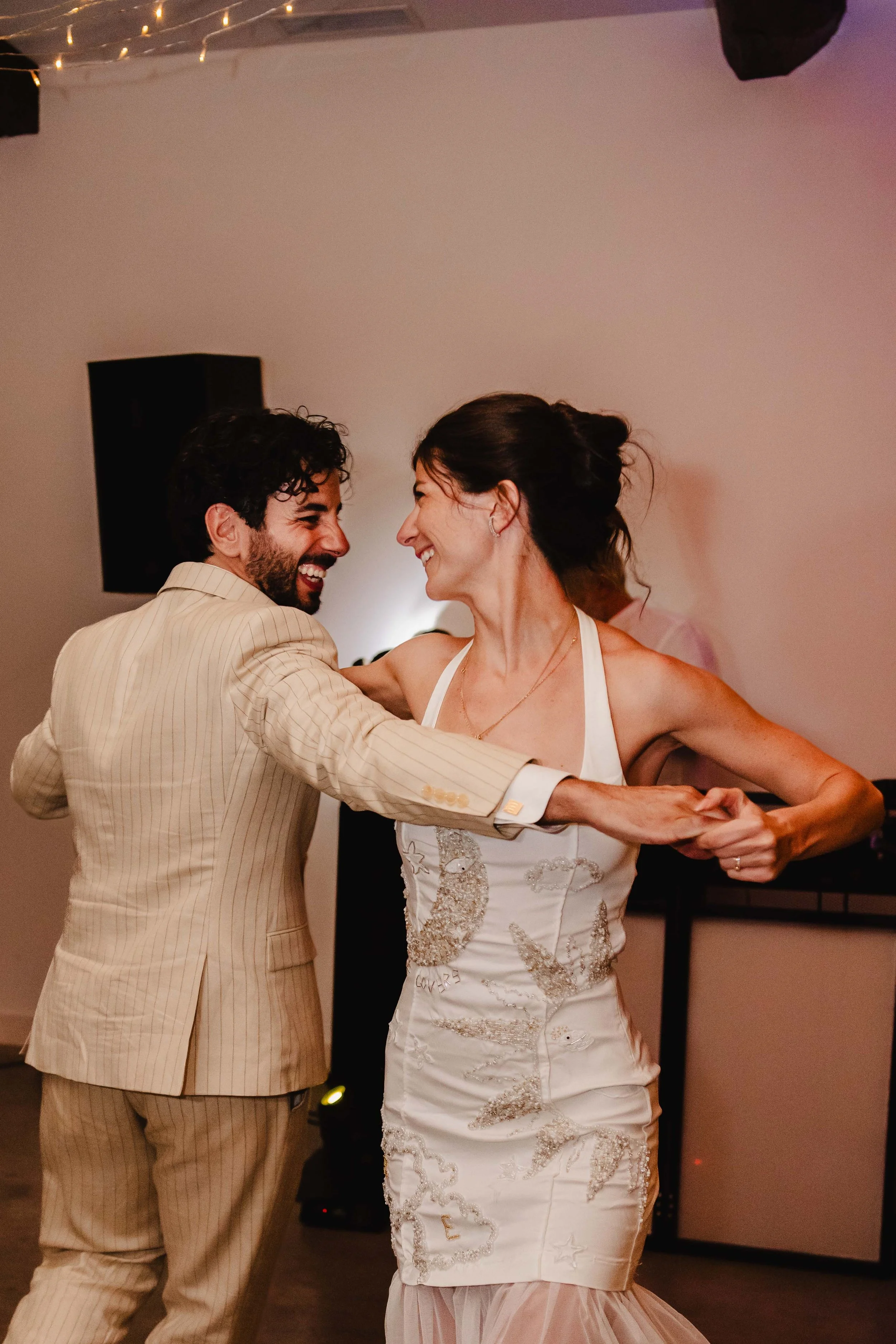 Un couple danse lors d'une fête, la femme porte une robe blanche avec des broderies dorées et l'homme un costume beige à rayures.