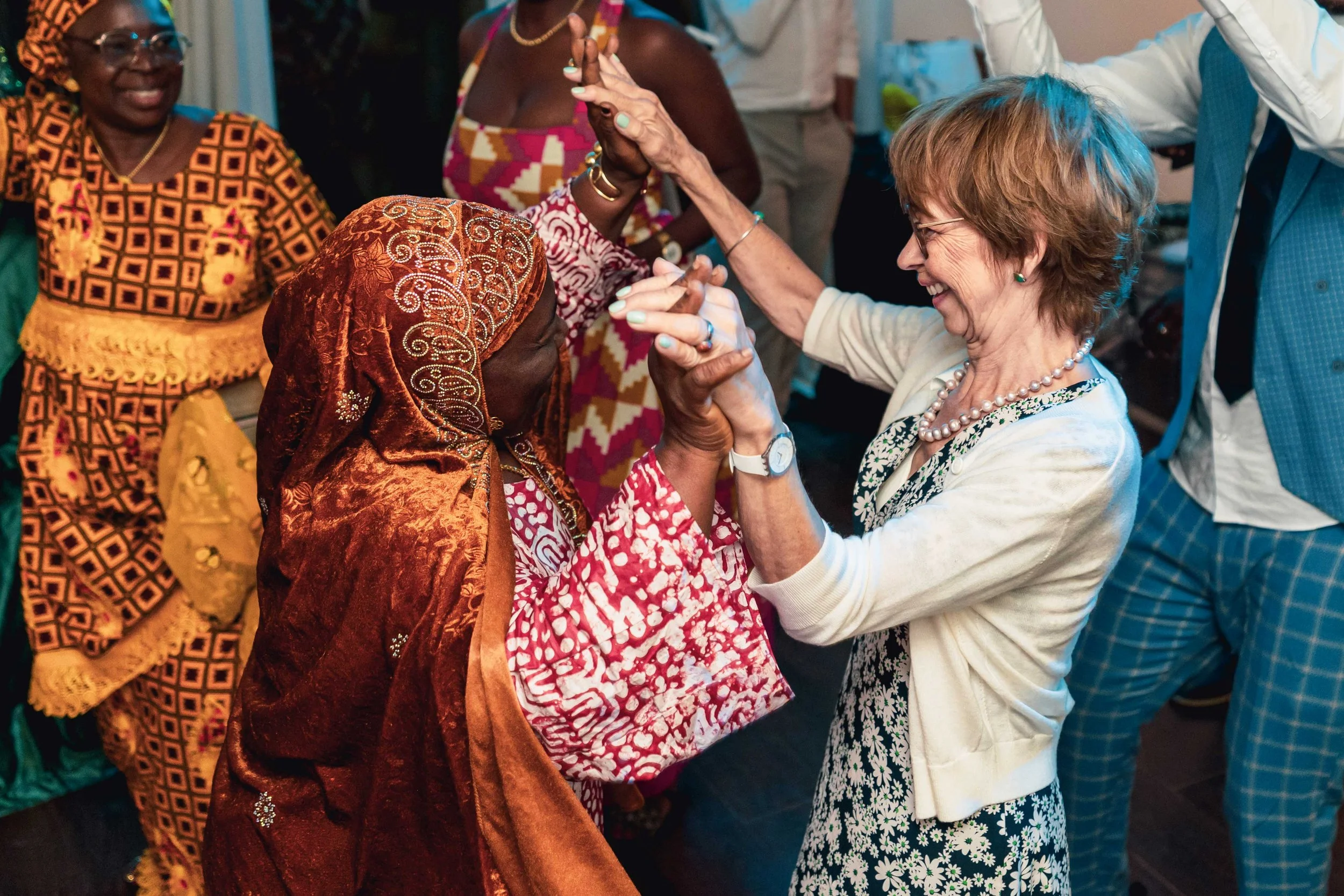 Deux femmes dansent et se tiennent par la main lors d'une célébration. L'une porte une robe colorée à motifs rouges et blancs avec un voile marron, l'autre porte un cardigan blanc avec un haut à motifs floraux noirs et blancs. D'autres personnes en t