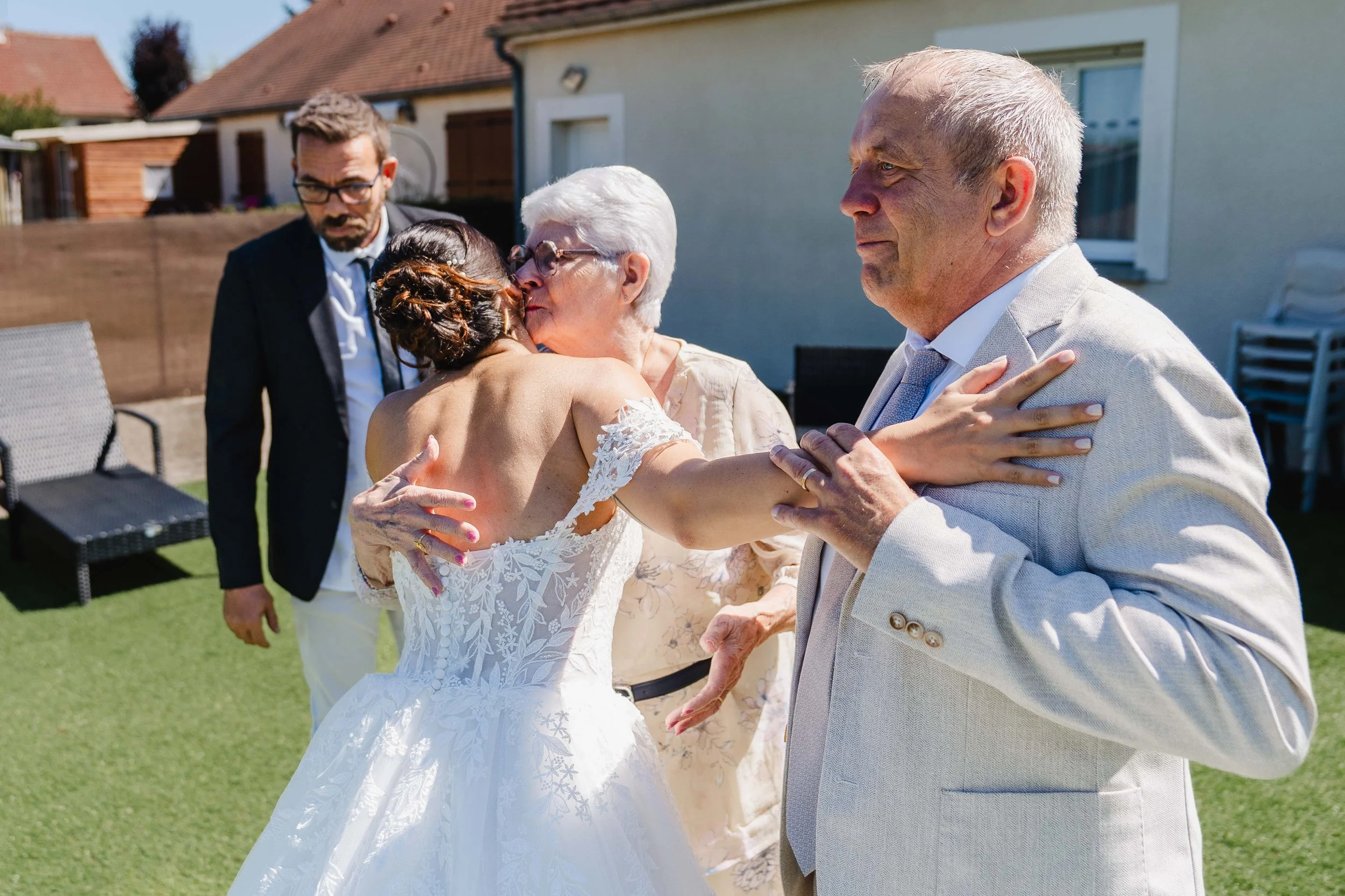Fête extérieure où une jeune femme en robe de mariée embrasse une femme âgée, entourée de deux hommes. Le soleil brille. 