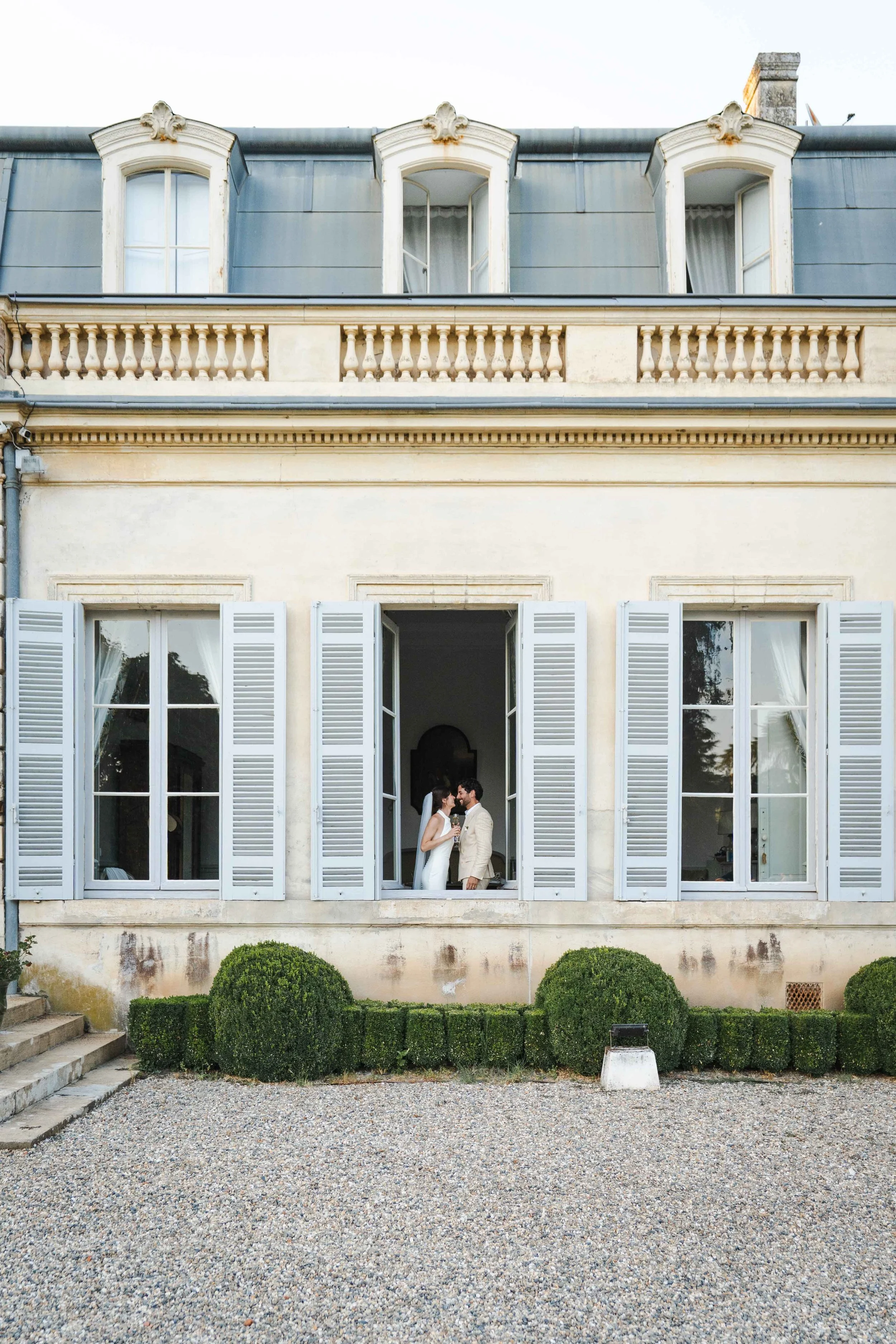 Un couple de mariés se tient à une fenêtre ouverte de la façade d'une maison élégante type parisienne, avec des volets en bois gris clair, dans un jardin avec des buissons taillés.
