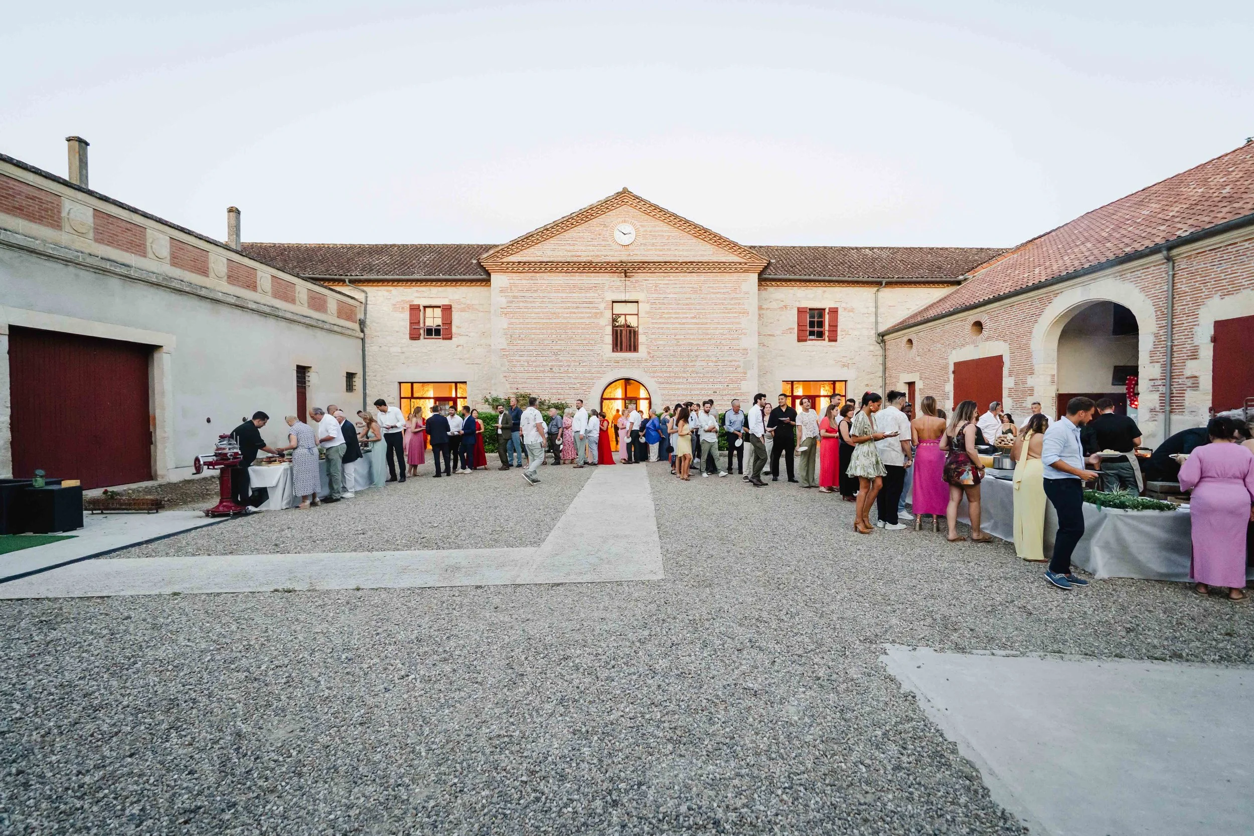 Un groupe de personnes en file d'attente lors d'une réception ou d'un événement à l'extérieur, devant le bâtiment en pierre du chateau Saint Denis, au crépuscule.
