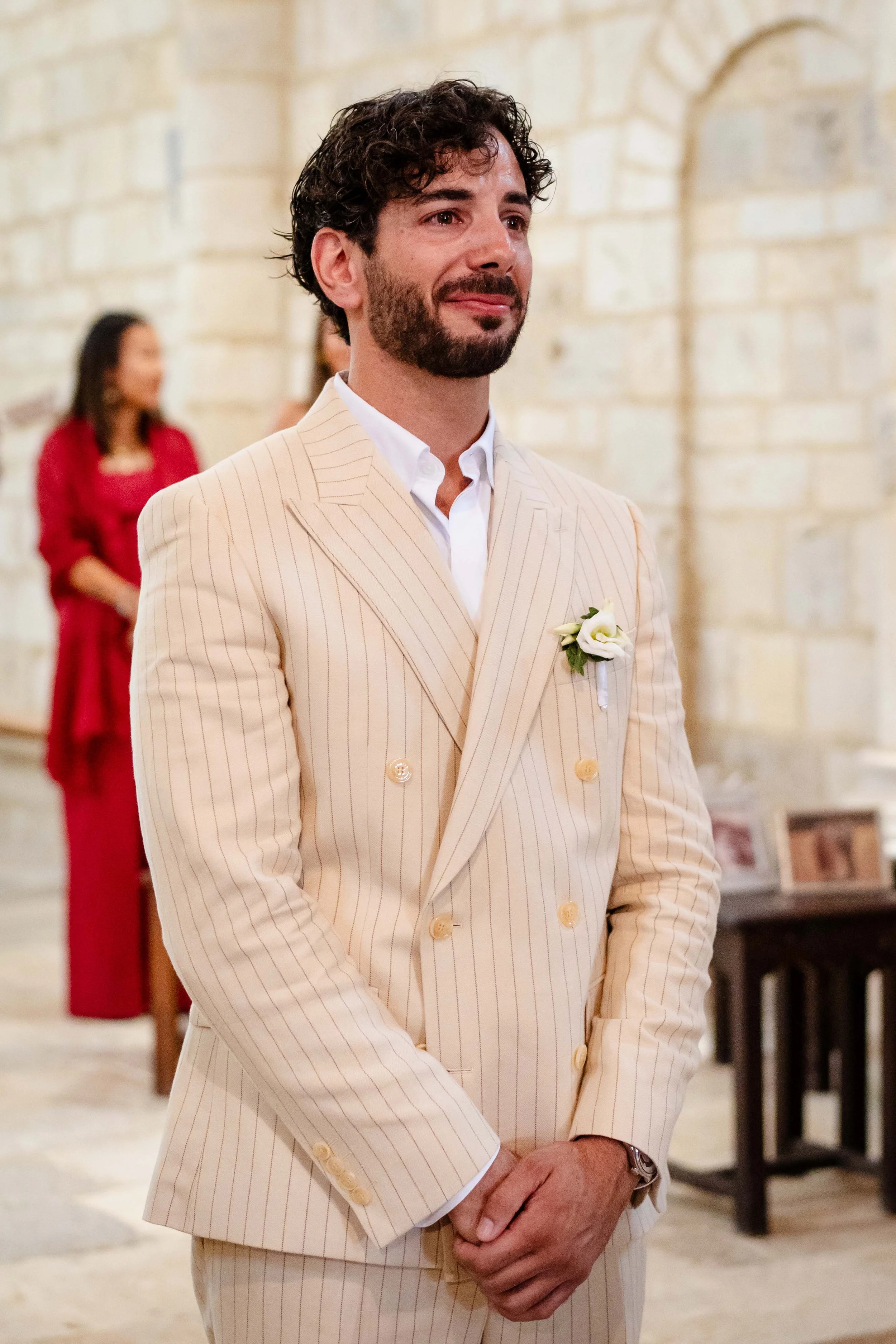 Un homme en costume beige à rayures, portant une boutonnière blanche, semble ému lors d'une cérémonie ou d'un mariage, avec des personnes en arrière-plan dans un lieu intérieur en pierre.