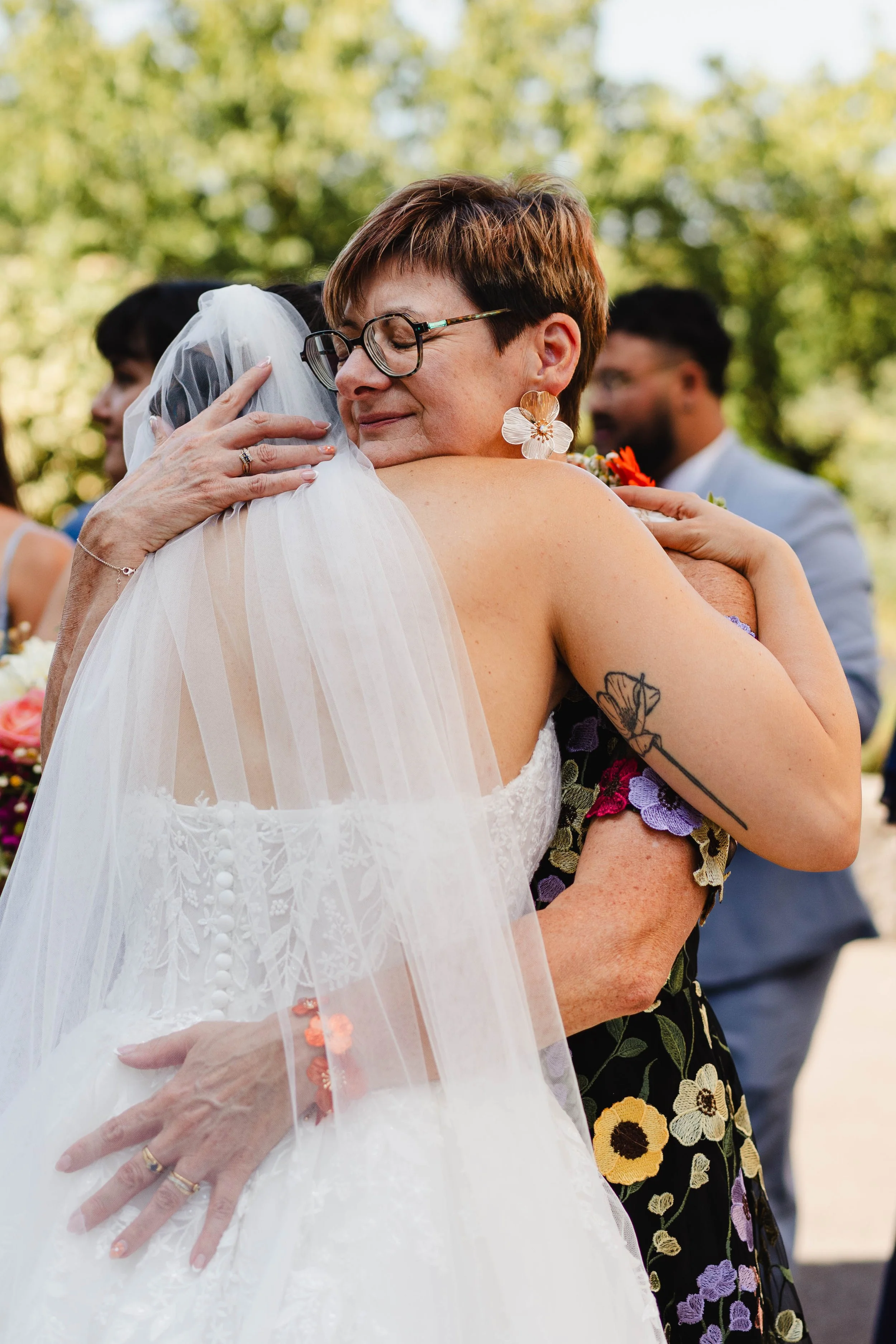 Deux femmes s'embrassent lors d'un mariage en extérieur, entourées d'autres personnes, avec des arbres en arrière-plan.