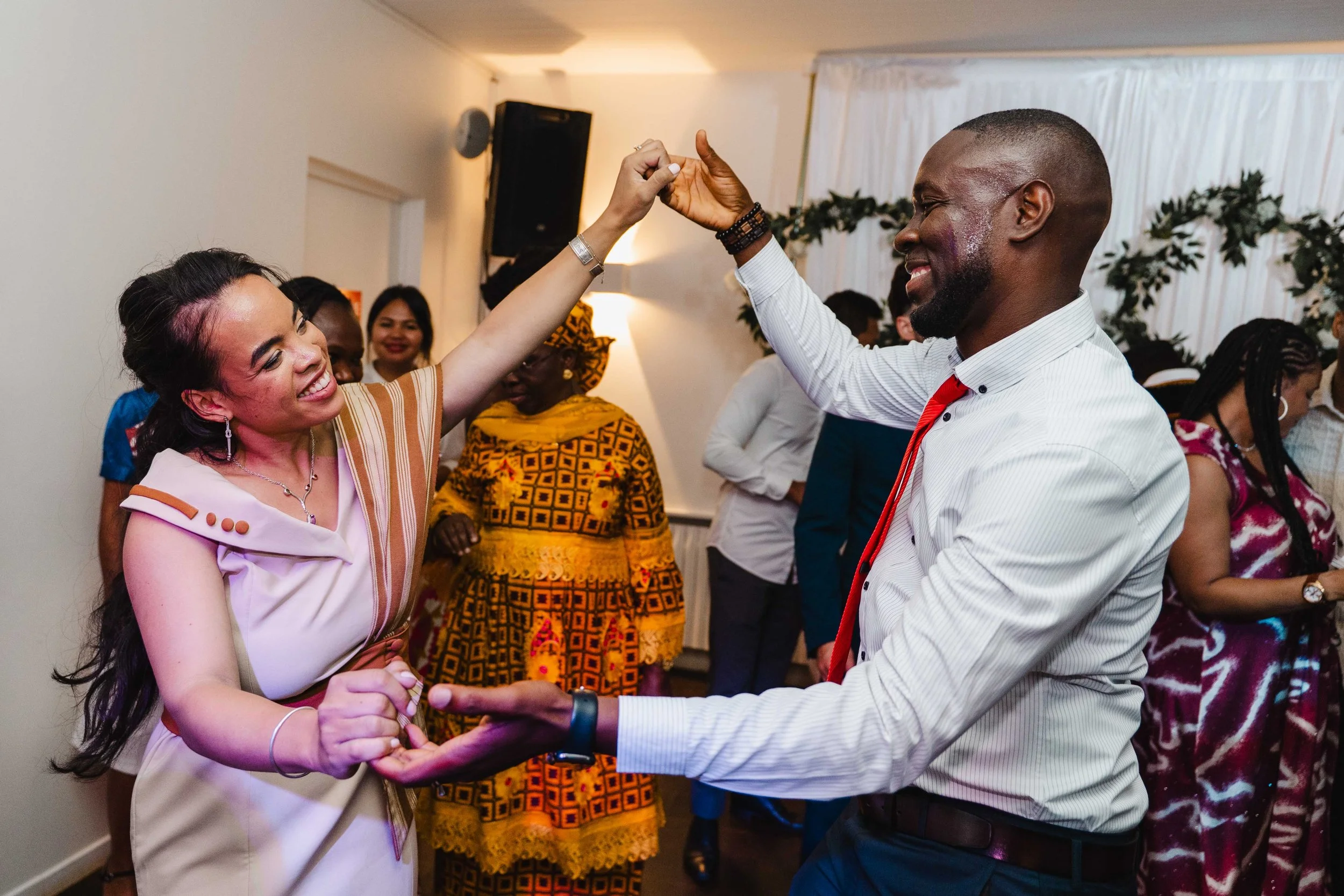 Un couple danse lors d'une fête, la femme sourit et l'homme la regarde avec bonheur, entourés d'autres personnes dans une salle décorée.