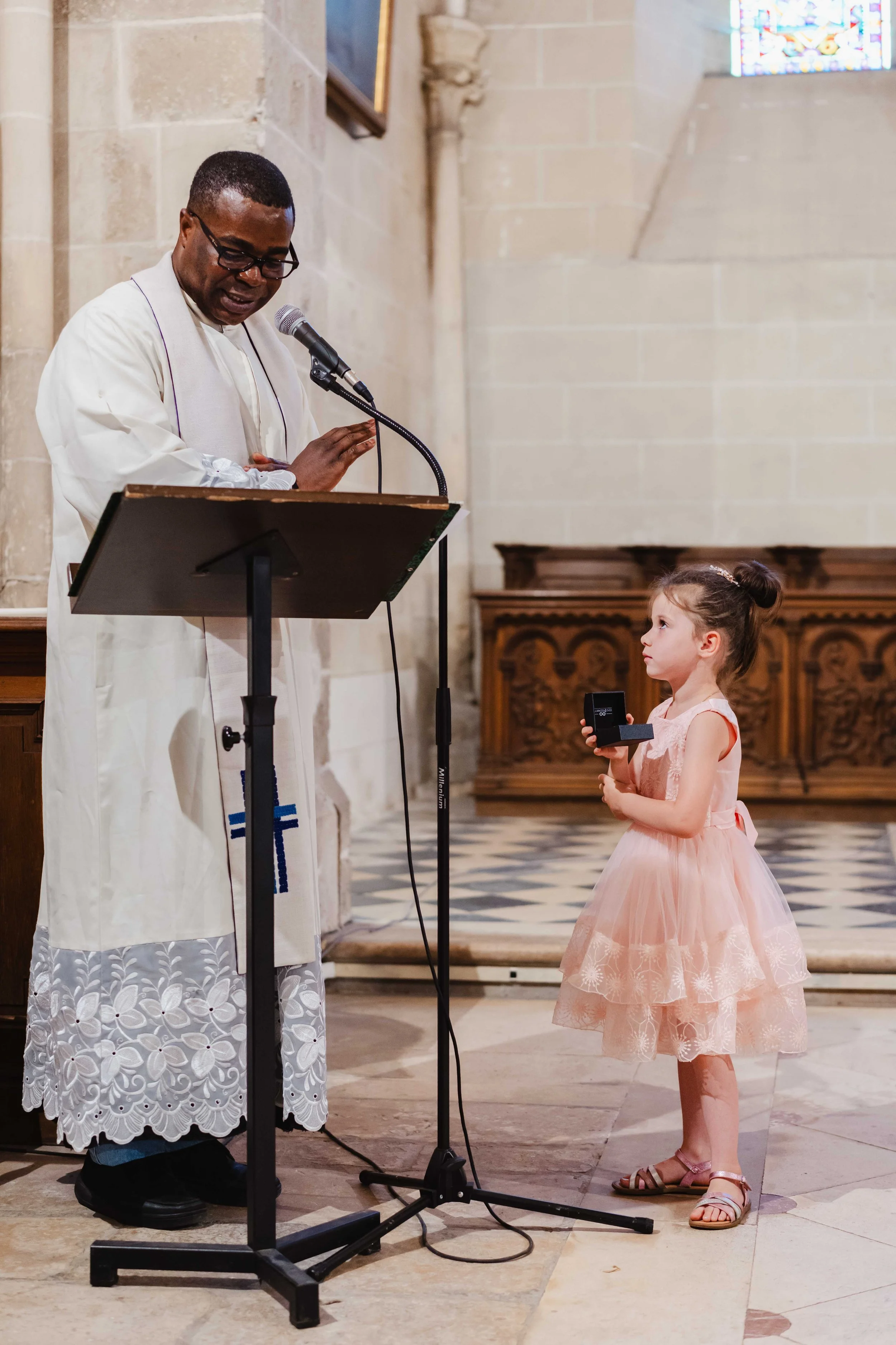 Un prêtre ou un officiant chantant ou parlant devant un micro dans une église, tandis qu'une jeune fille en robe de cérémonie assortie, tenant une petite boîte, regarde attentivement.
