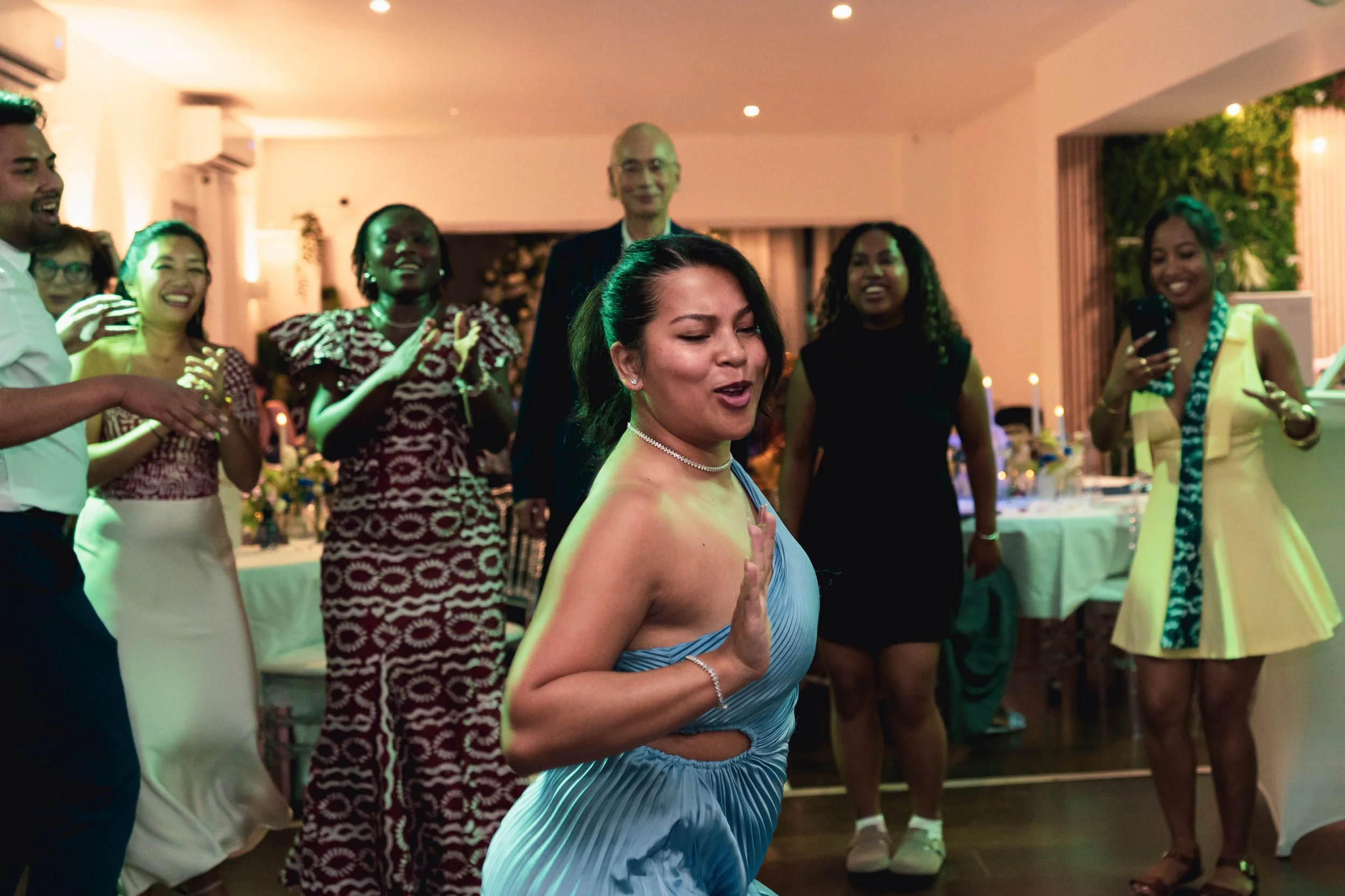 Un groupe de personnes dansant et souriant lors d'une fête dans une salle éclairée, vêtements festifs, ambiance joyeuse.