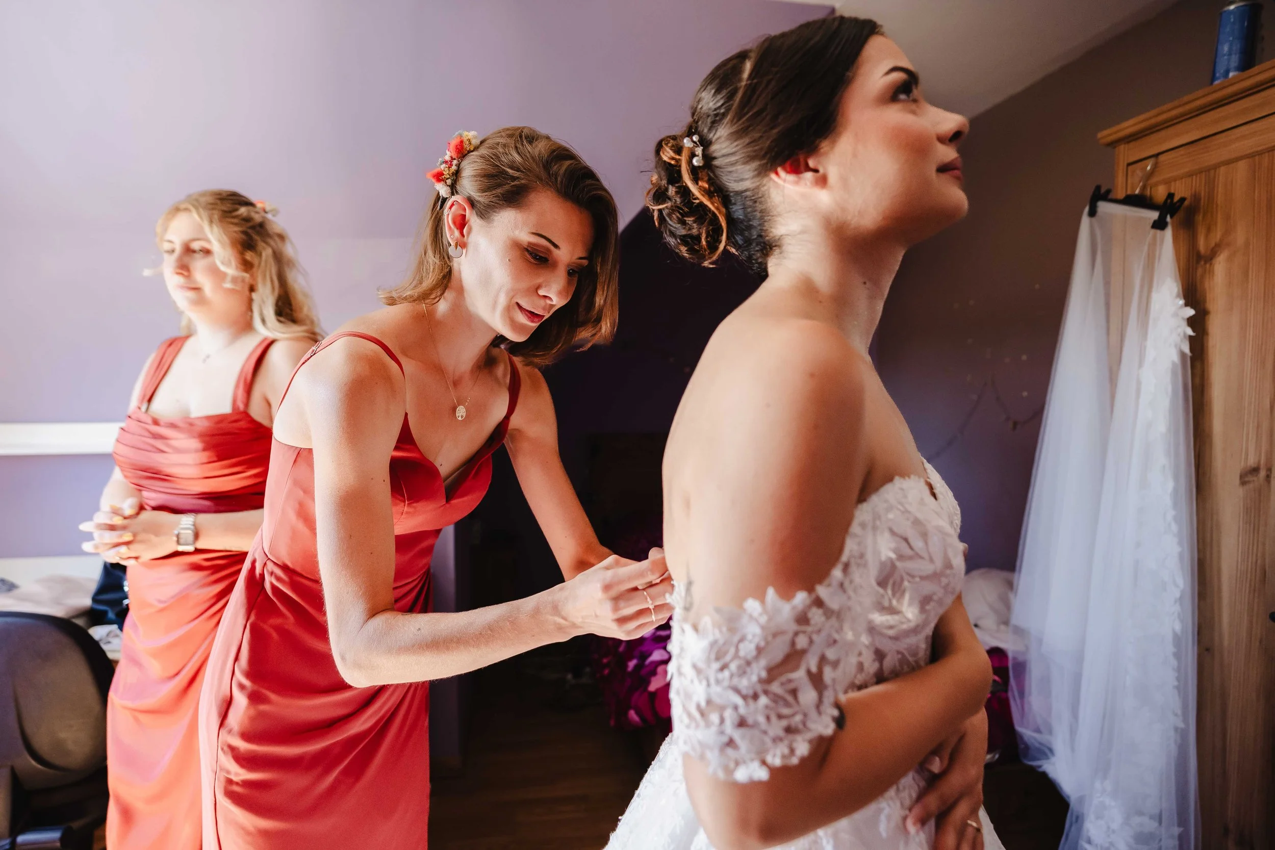 Une femme en robe de mariée se fait aider à boutonner par une autre femme portant une robe rouge, dans une pièce où une troisième femme en robe rouge est en arrière-plan.