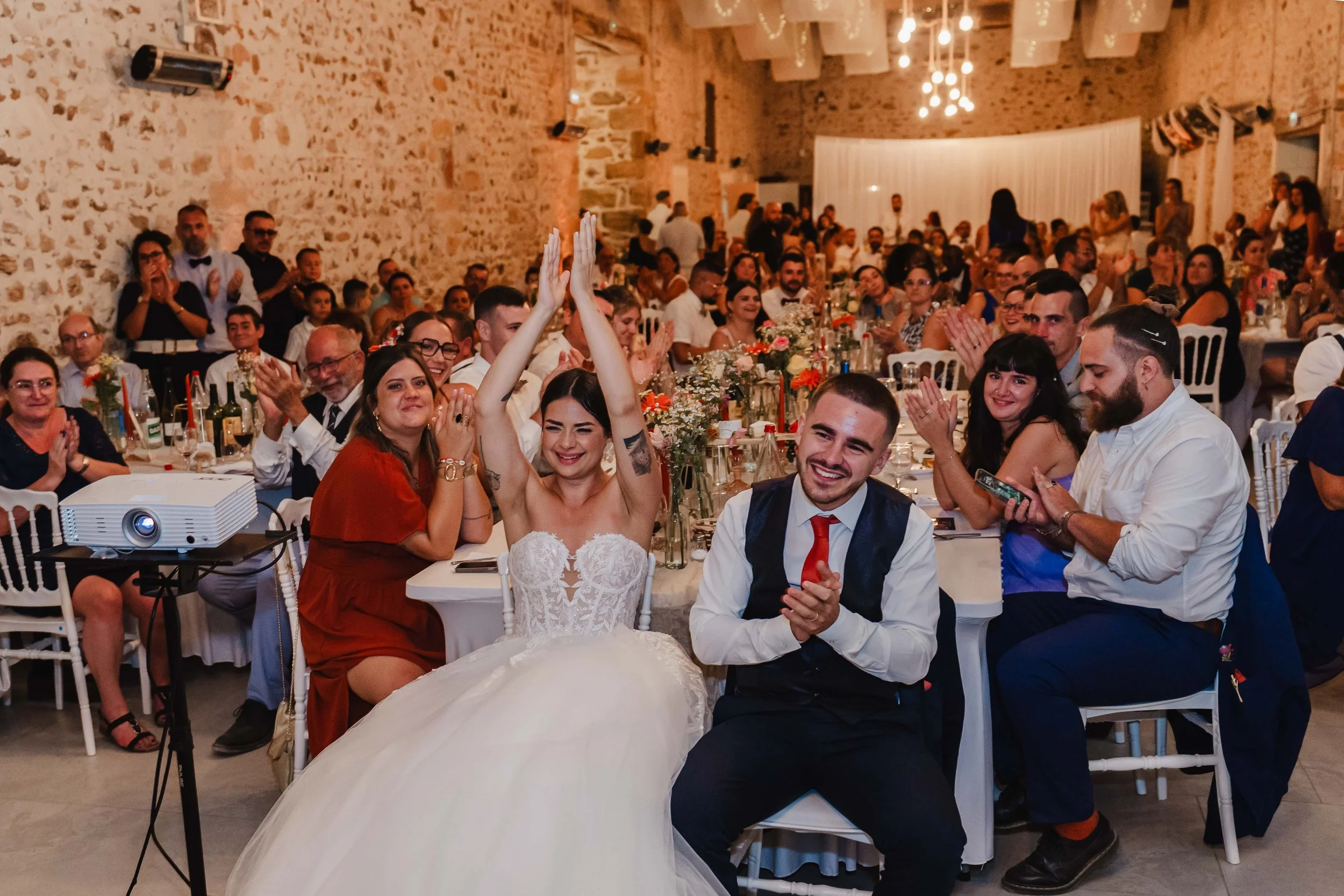 Moment de fête à un mariage, avec le couple principal qui rit et lève les mains en triomphe, entouré de nombreux invités assis à une grande table décorée de fleurs et de bouteilles, dans une salle au mur en pierre, éclairée par un éclairage chaleureu