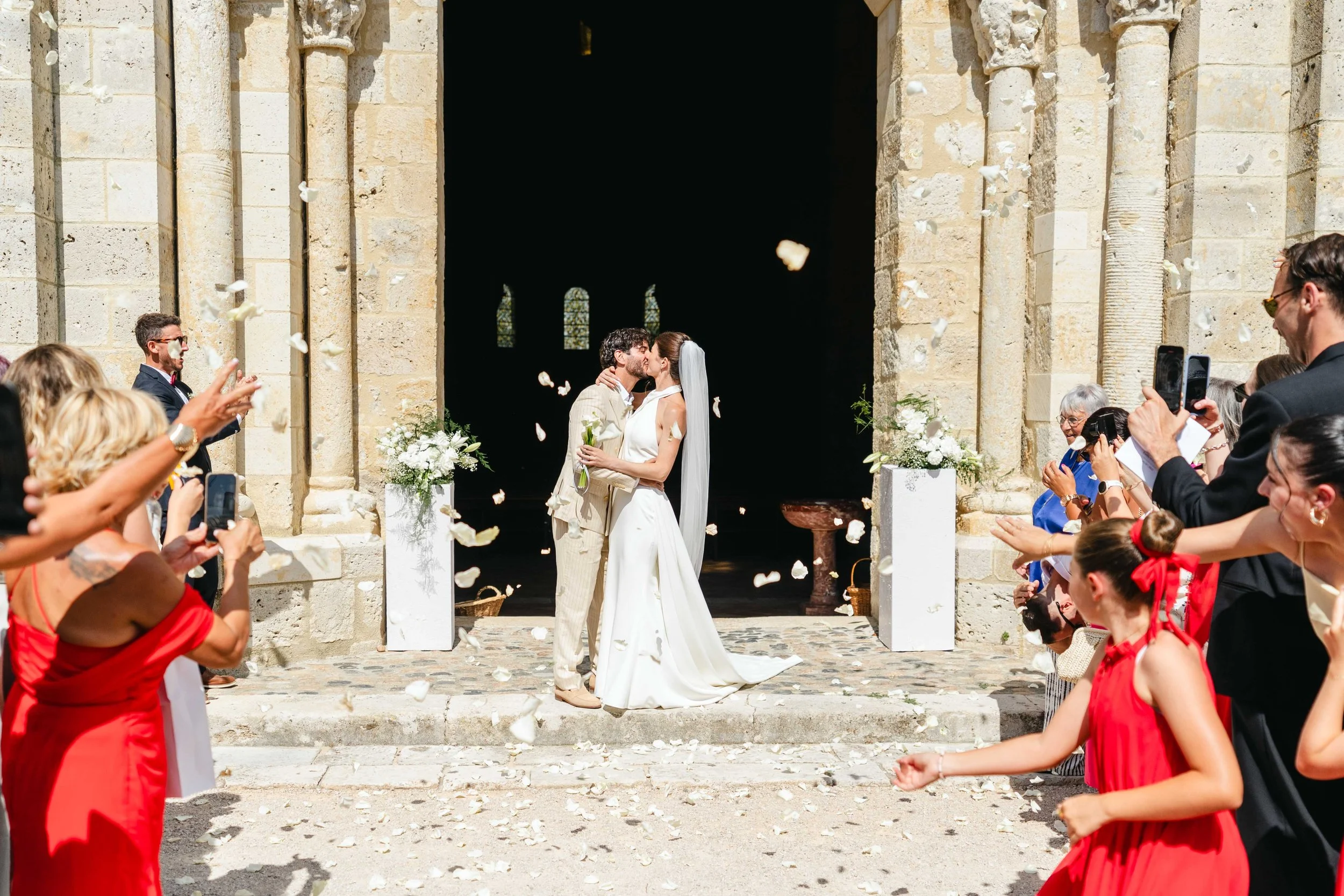 Un couple de mariés s'embrassent devant l'entrée de l'église de Moirax, entourés d'invités jetant des pétales de fleurs, célébrant leur mariage lors d'une journée ensoleillée.
