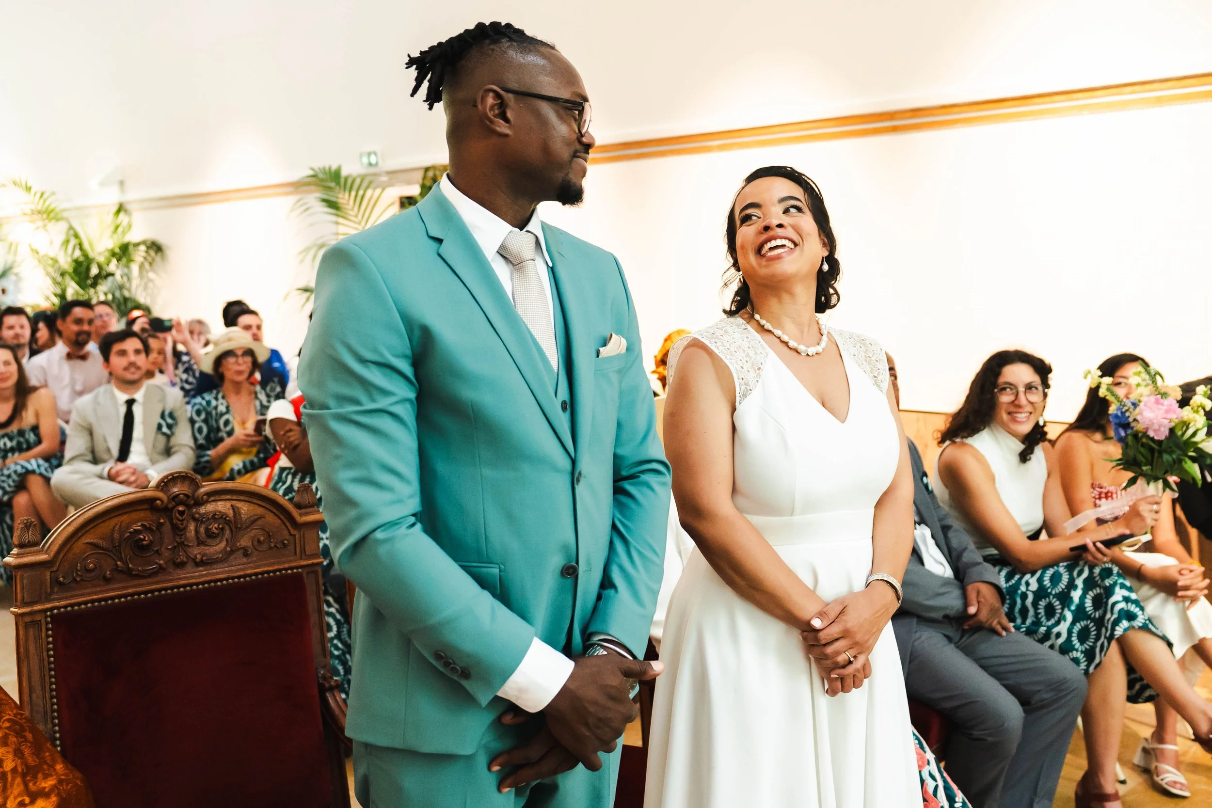 Deux personnes lors d'une cérémonie, une femme en robe blanche souriante et un homme en costume turquoise, entourées de plusieurs invités assis.