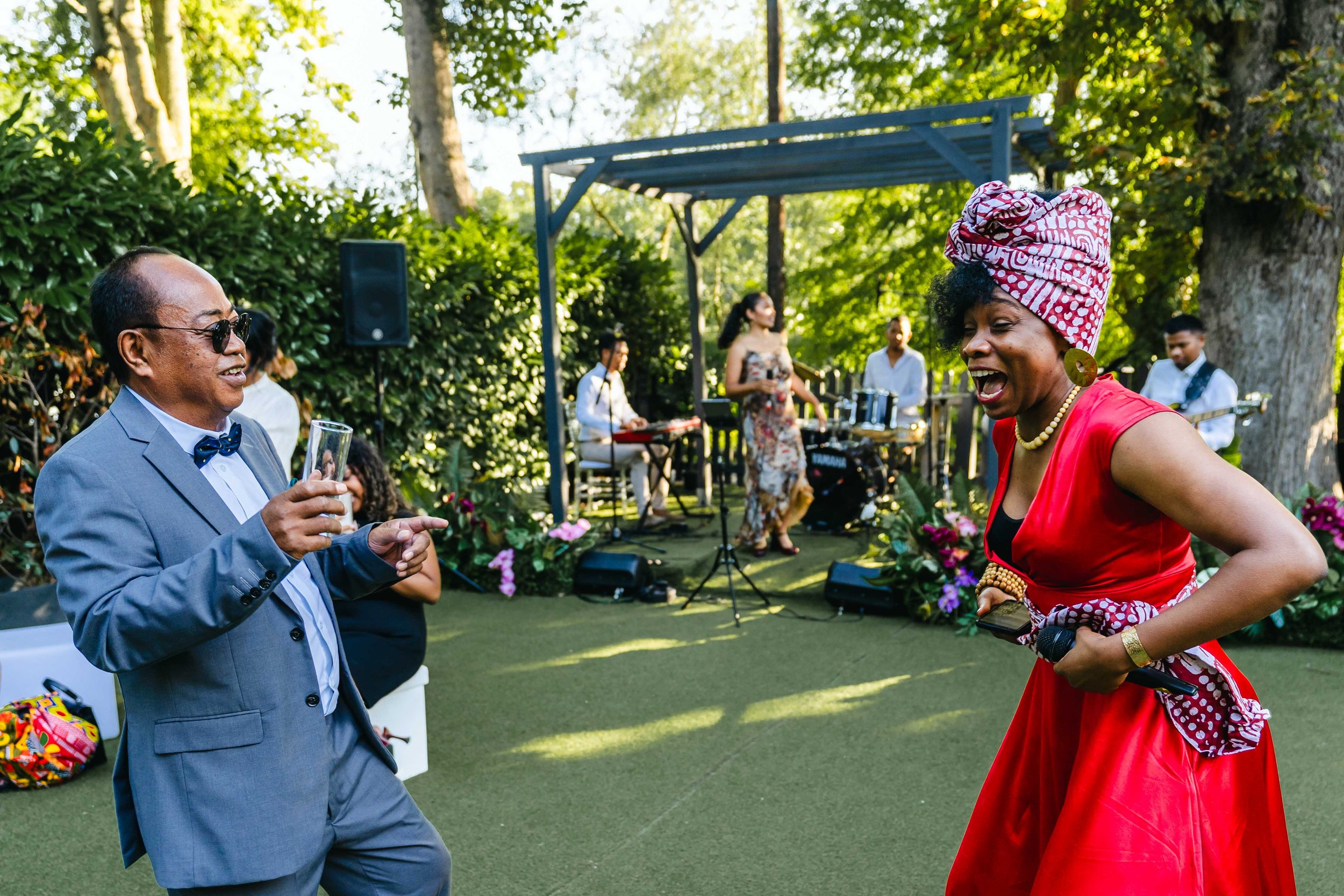 Deux personnes dansent et rient lors d'une fête en plein air avec un groupe de musiciens en arrière-plan. L'une porte un costume gris avec un nœud papillon, l'autre une robe rouge avec un headwrap rouge et blanc.