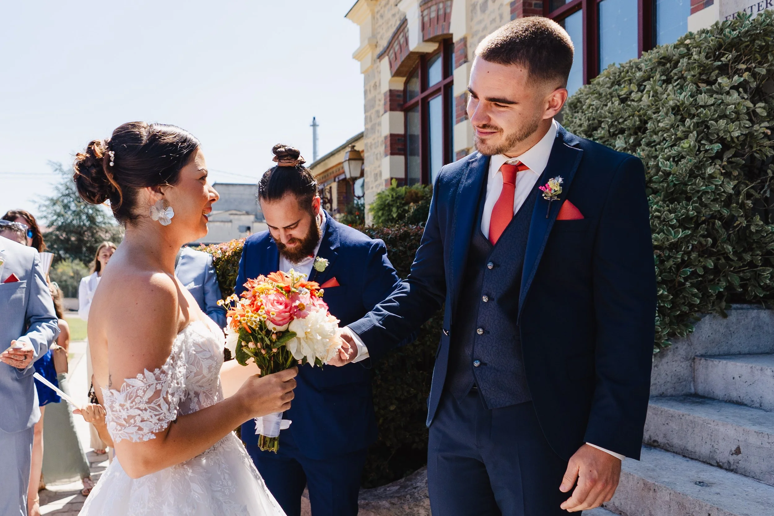 Un mariage avec un homme en costume élégant, une femme en robe blanche tenant un bouquet de fleurs, entourés de proches sous un ciel ensoleillé devant un bâtiment historique.