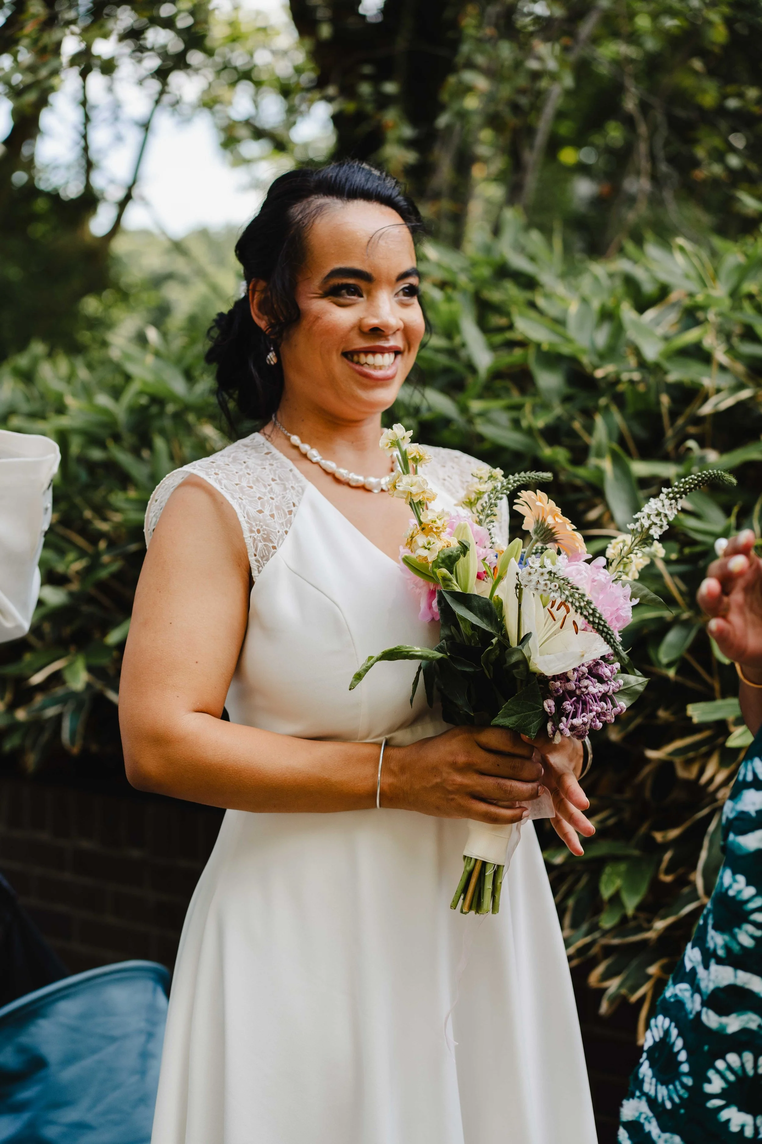 Une femme souriante en robe blanche avec un bouquet de fleurs, lors d'une cérémonie en plein air entourée de verdure.