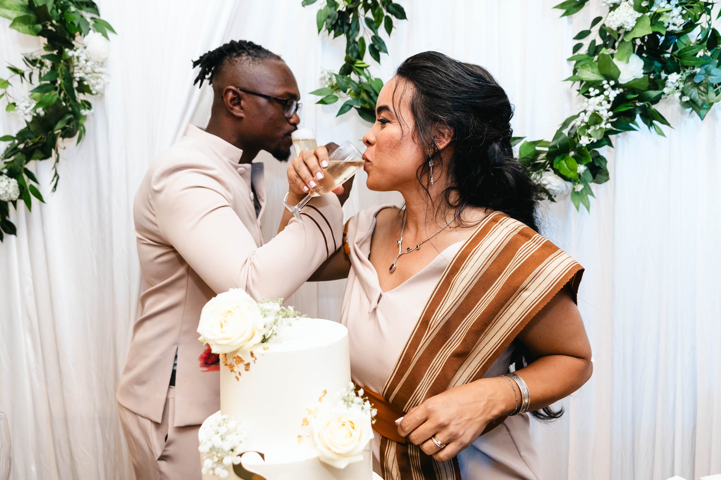 Deux personnes à un mariage, la femme boit du champagne tandis que l'homme lui donne la coupe, avec un gâteau blanc décoré de fleurs à l'avant.