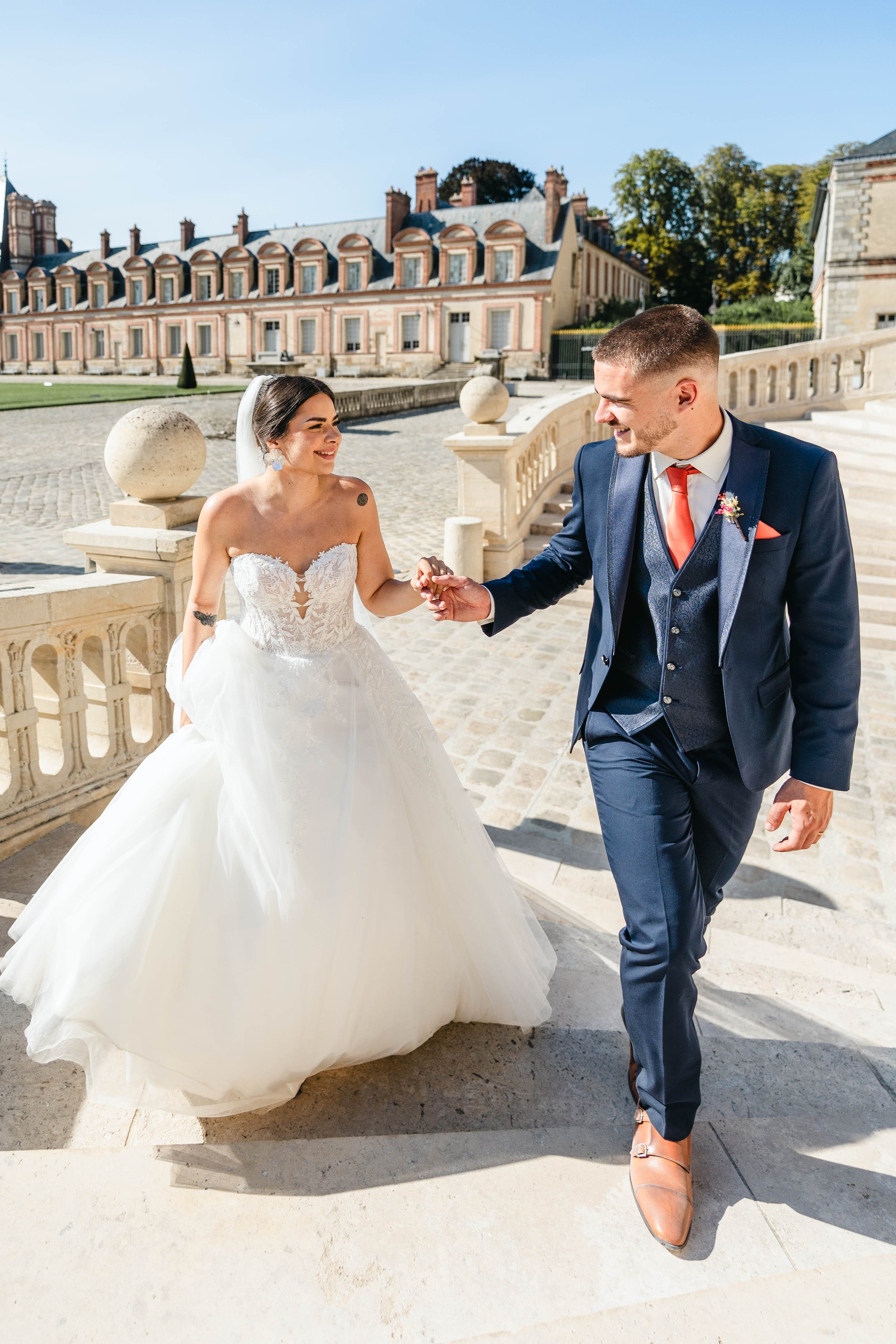 Un couple de mariés, un homme et une femme en robe de mariage, se tenant la main et souriant, devant le château de Fontainebleau. Le marié porte un costume bleu avec une cravate rouge, et la mariée porte une robe blanche avec un voile.