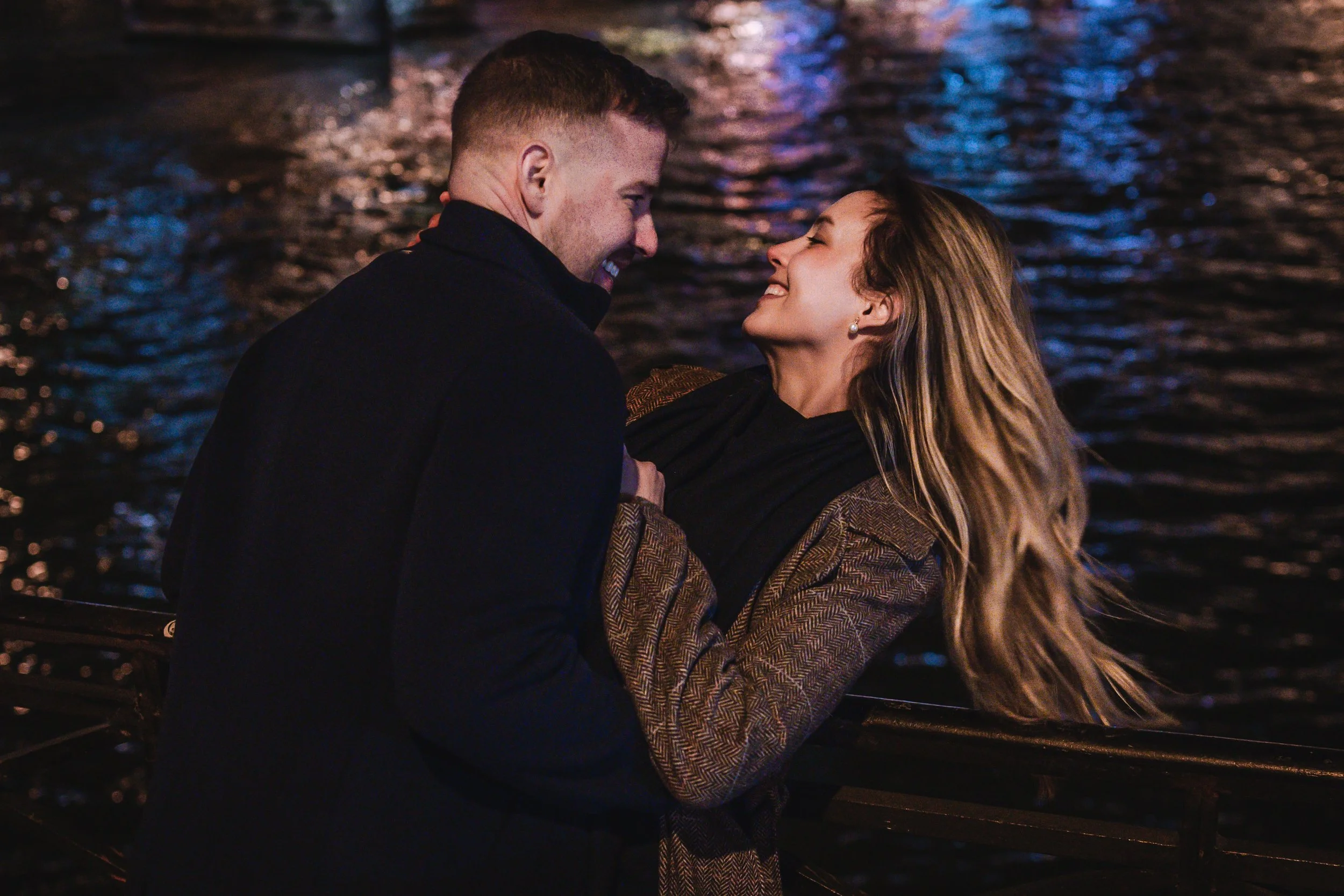 Un couple souriant, en tenue élégante, se tenant face à face près de la Seine à paris, sous un éclairage doux et coloré.