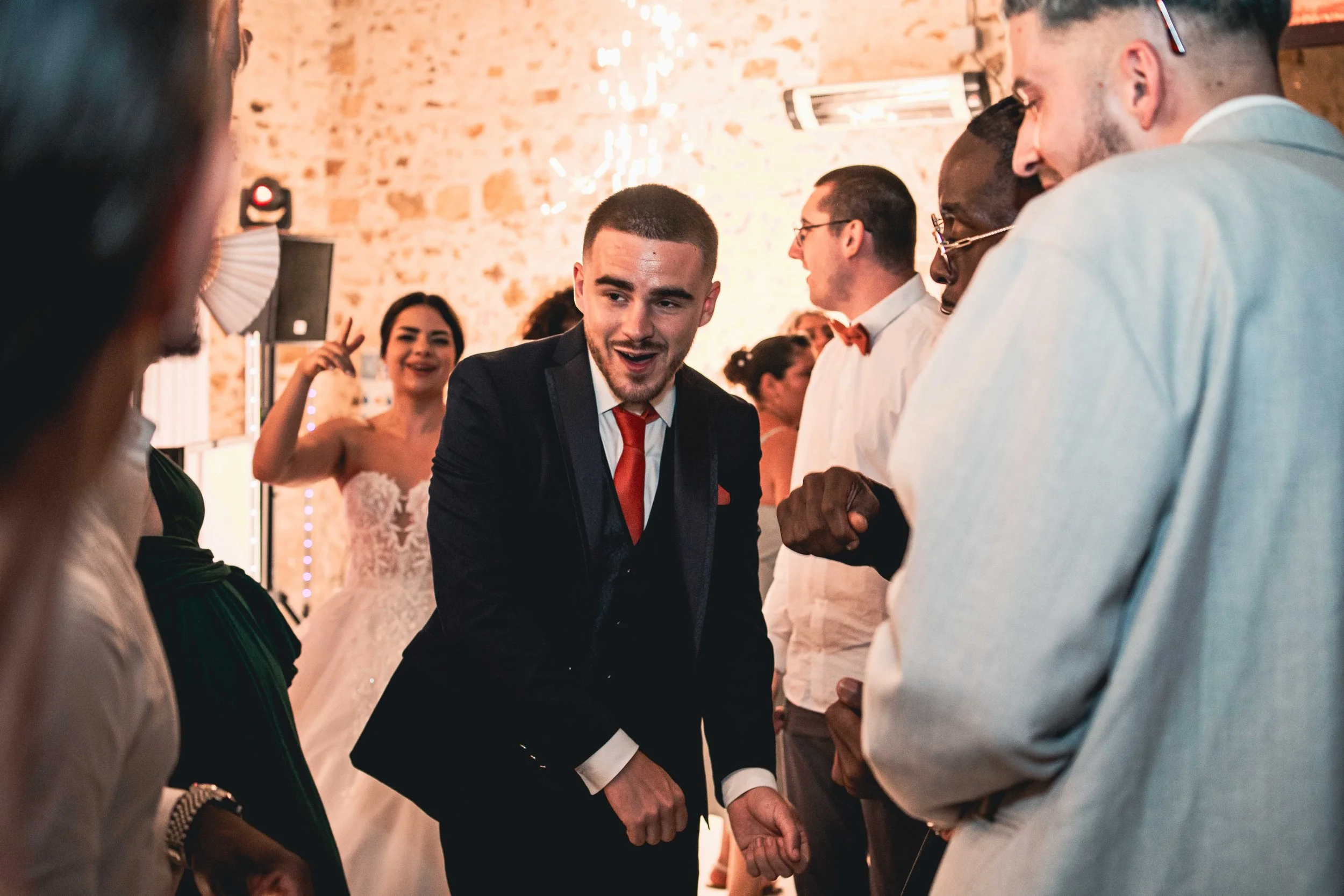 Groupe de personnes dansant et s'amusant lors d'une fête ou d'une célébration, avec un homme en costume noir et rouge au centre, entouré de plusieurs autres personnes.