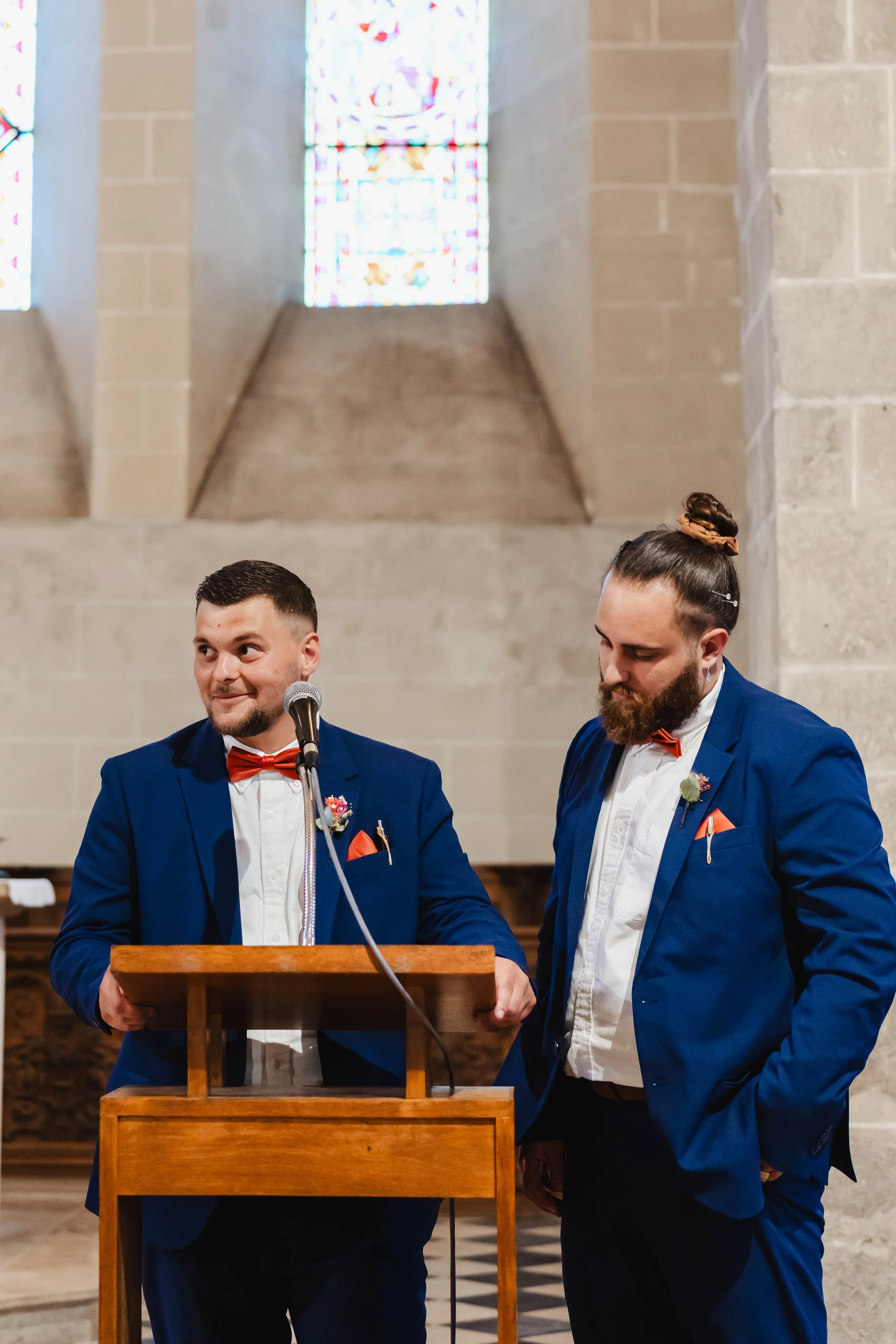 Deux hommes en costume bleu avec des nœuds papillons rouges, debout à une chaire en bois à l'intérieur d'une église, avec un vitrail coloré derrière eux.