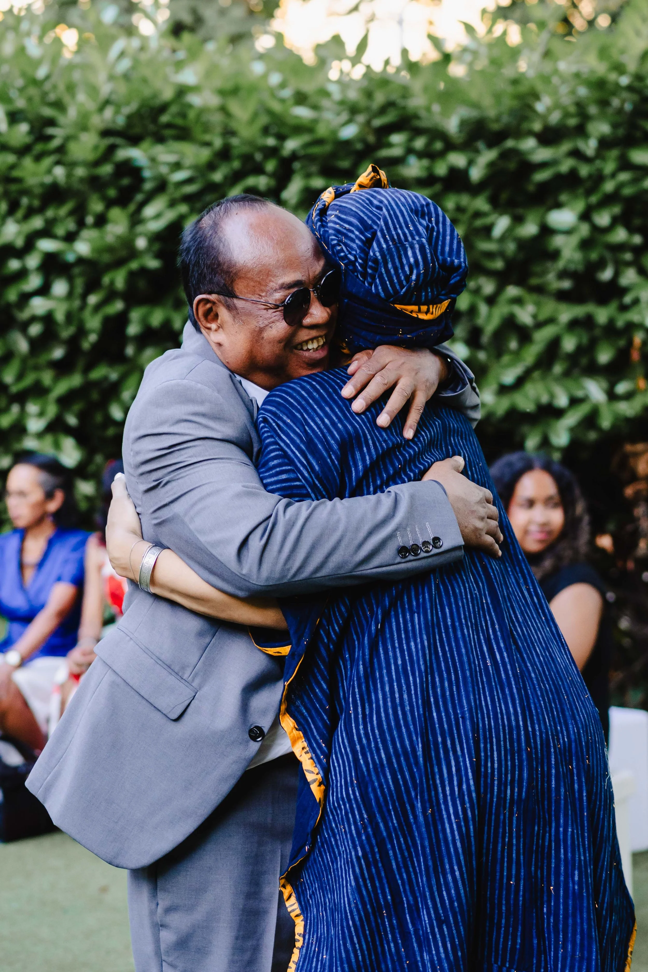 Deux personnes s'embrassent lors d'une rencontre émouvante en extérieur, avec un groupe d'autres personnes assises en arrière-plan, sous un arbre.