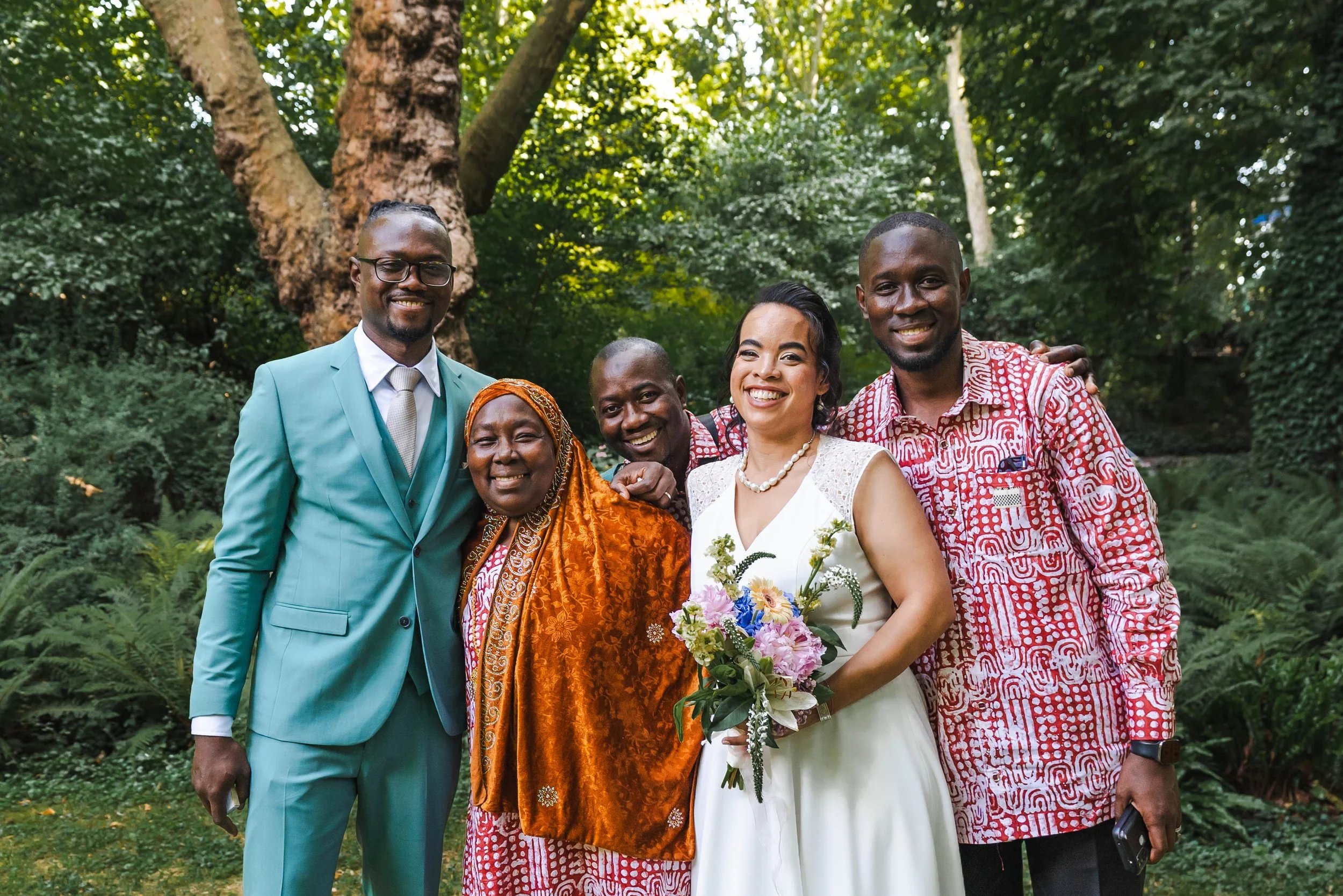 Groupe de personnes souriantes lors d'une occasion spéciale dans un espace vert, avec une femme portant une robe blanche et tenant un bouquet de fleurs, entourée de quatre hommes et une femme dans des vêtements colorés.