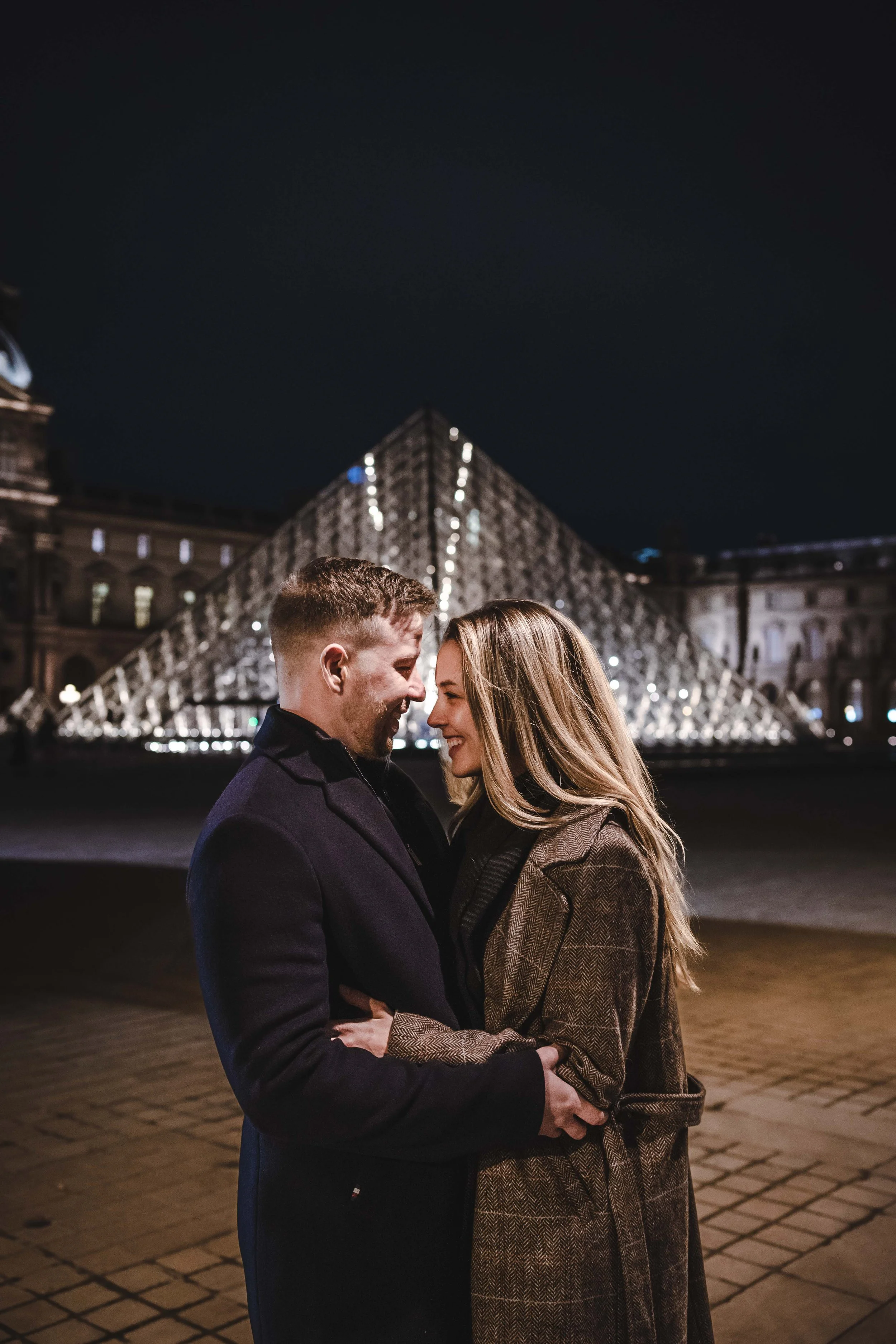 Un couple souriant et s'embrassant devant le musée du Louvre illuminé à Paris la nuit.