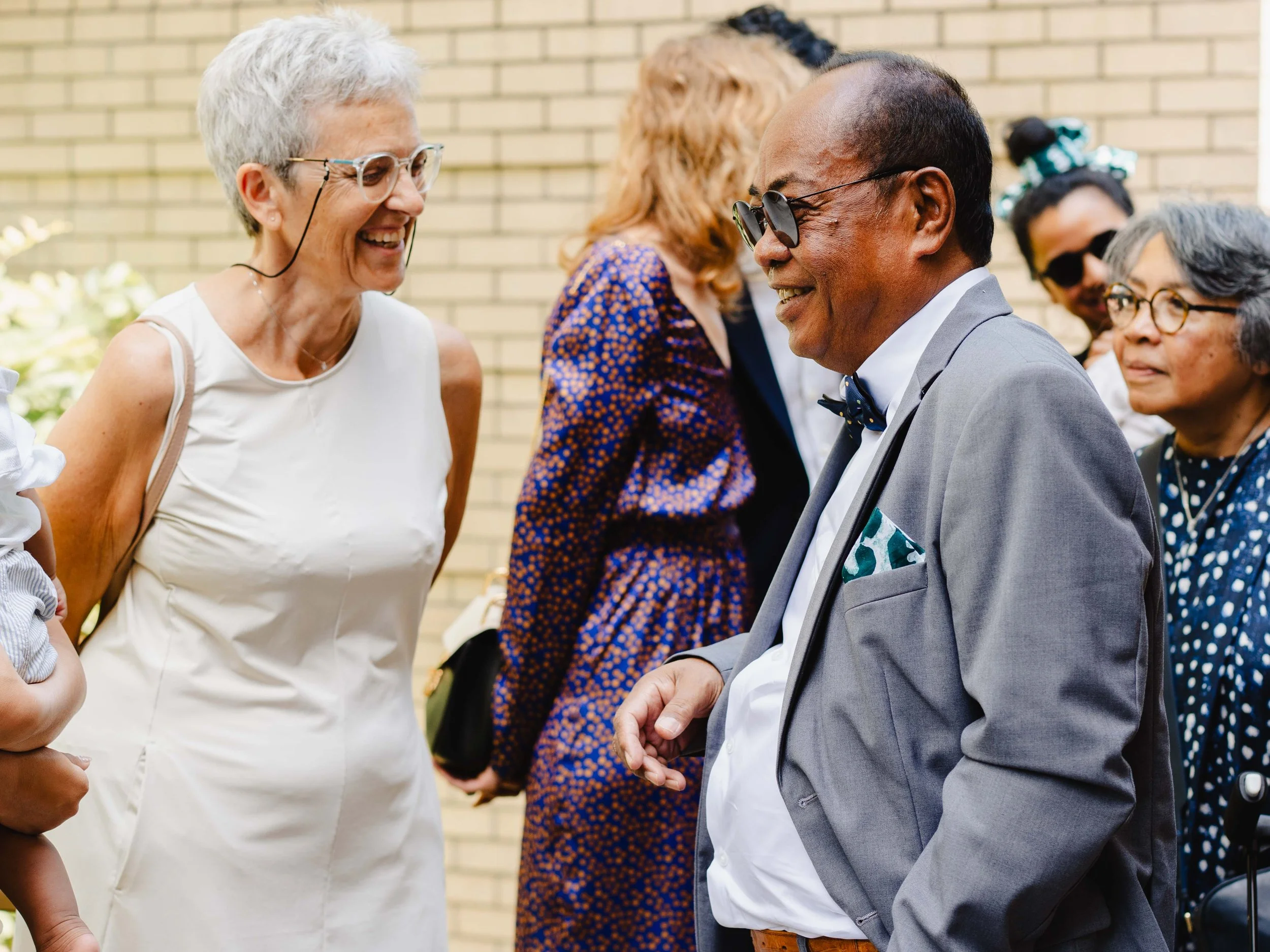 Un groupe de personnes âgées souriantes et déguisées, en train de converser lors d'une fête en extérieur, avec un fond de mur en briques.