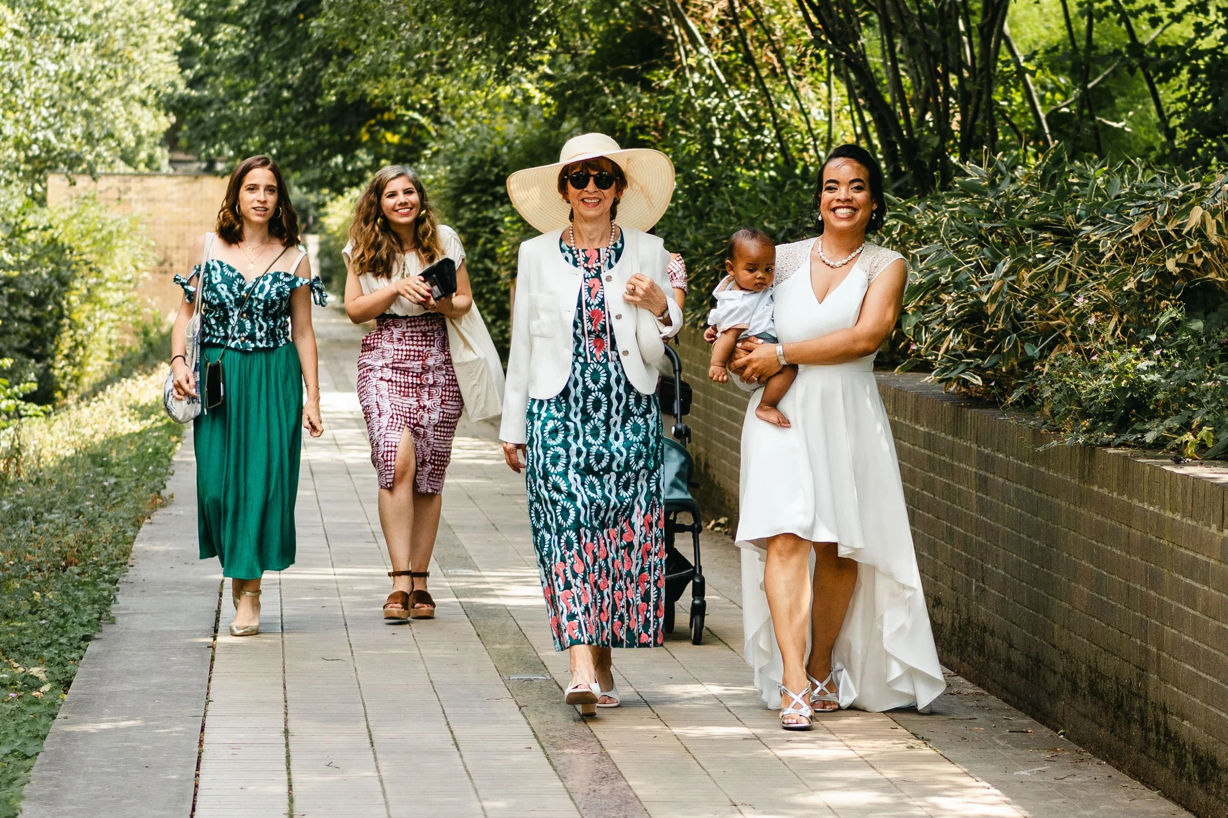 Groupe de cinq femmes marchant dans un parc, deux sont accompagnées d'un bébé, tous souriants et habillés élégamment, entourés de verdure