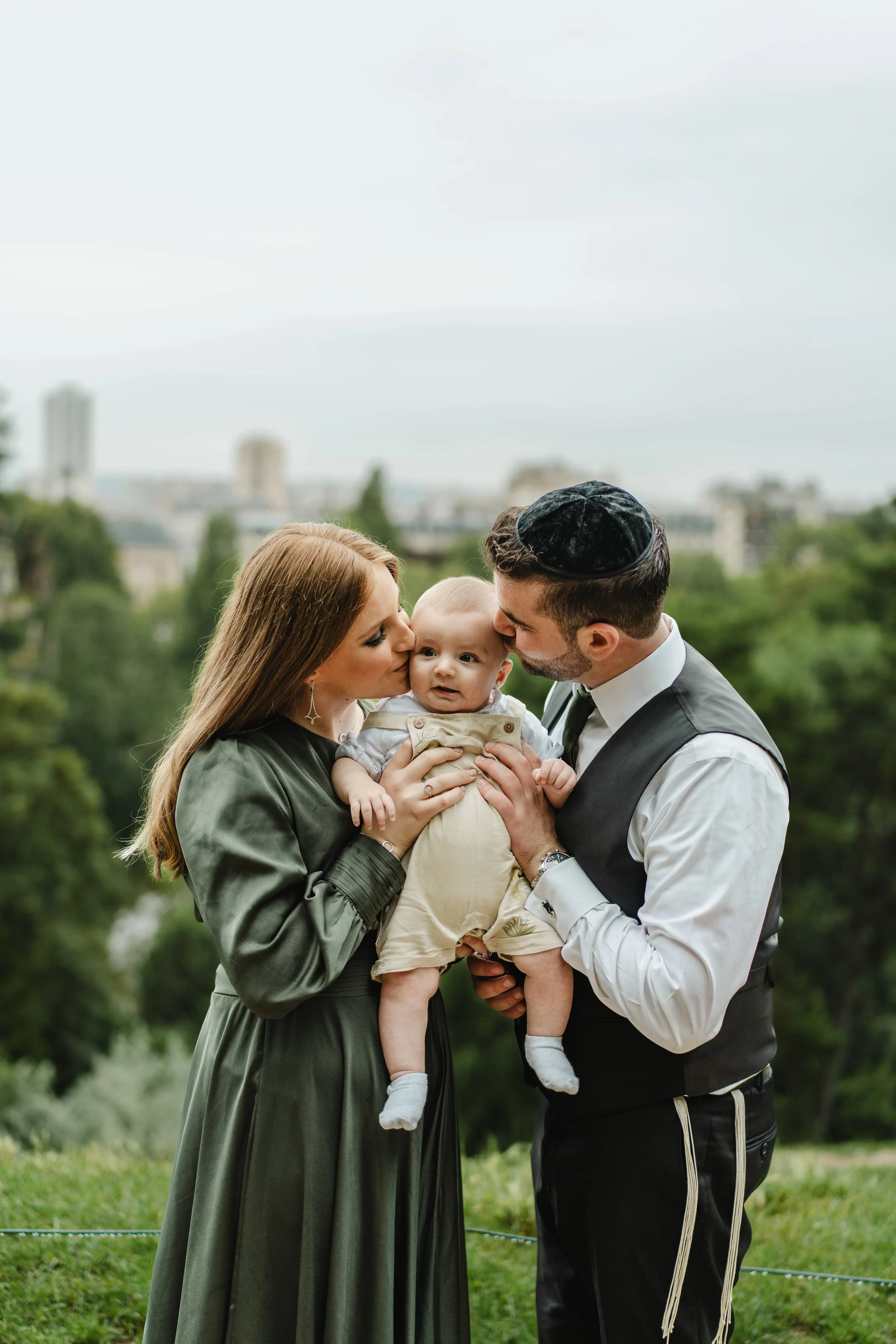 Une famille de trois personnes, un homme, une femme et un bébé, lors d'une cérémonie religieuse juive, l'homme porte un kippah et la femme porte une robe longue verte foncée. La famille s'embrasse et échange des baisers, avec un fond de nature et une