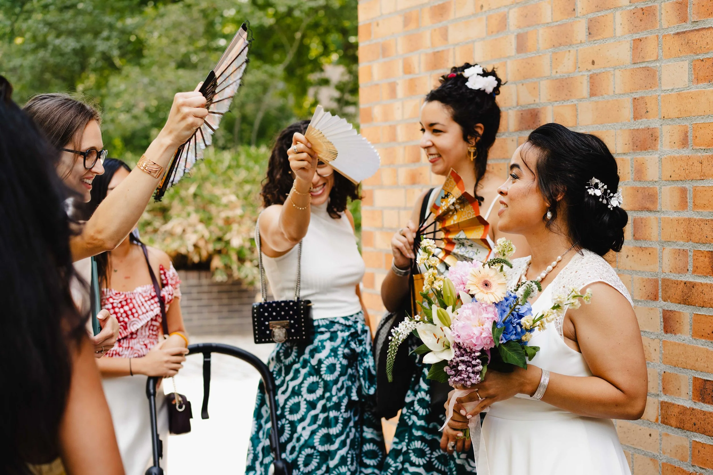 Femme en robe blanche tenant un bouquet de fleurs, entourée de femmes souriantes avec des éventails, en extérieur près d'un mur en briques.