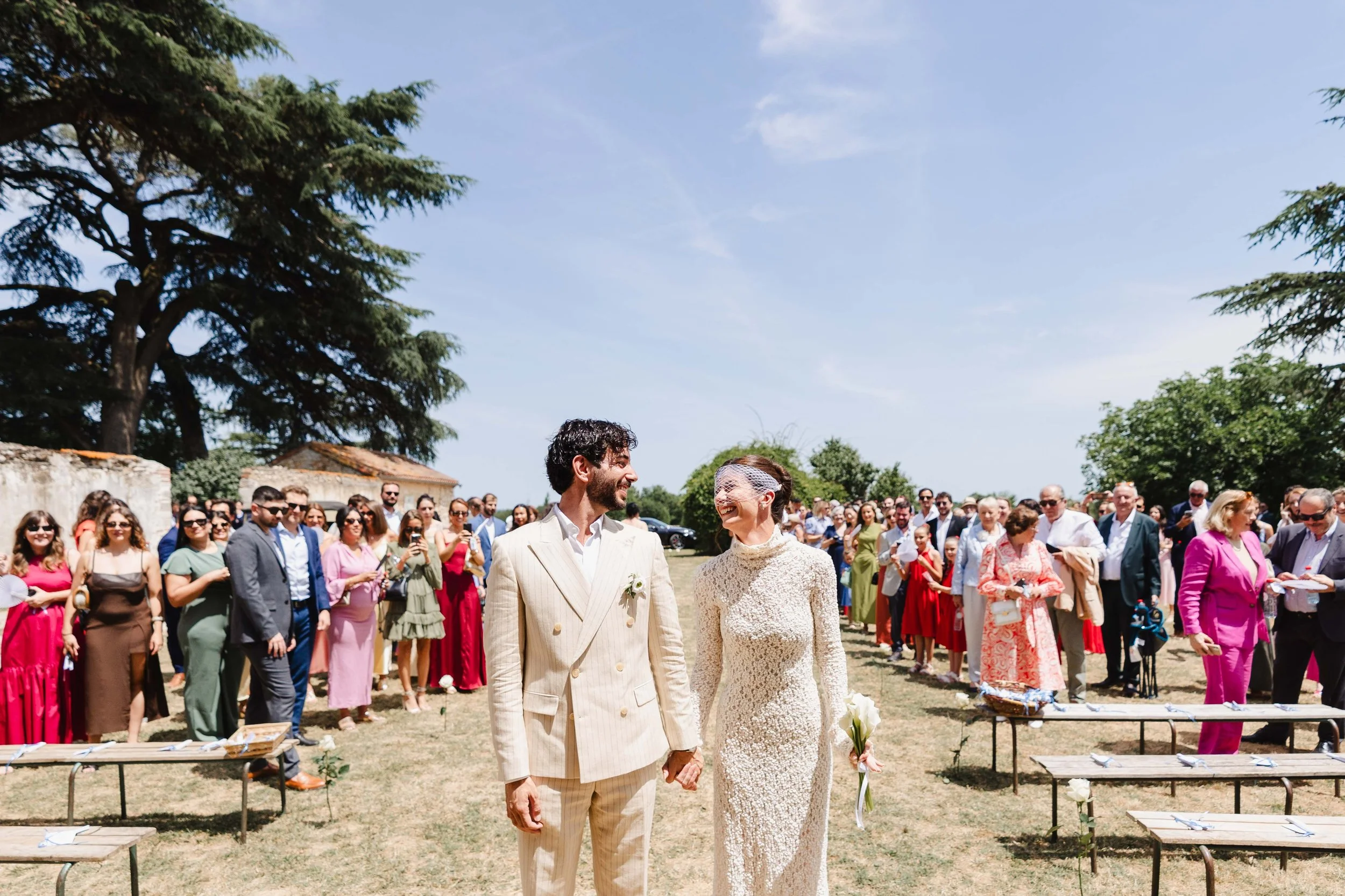 Un couple marié se tenant la main lors d'une cérémonie en plein air, entouré de nombreux invités sous un ciel ensoleillé.