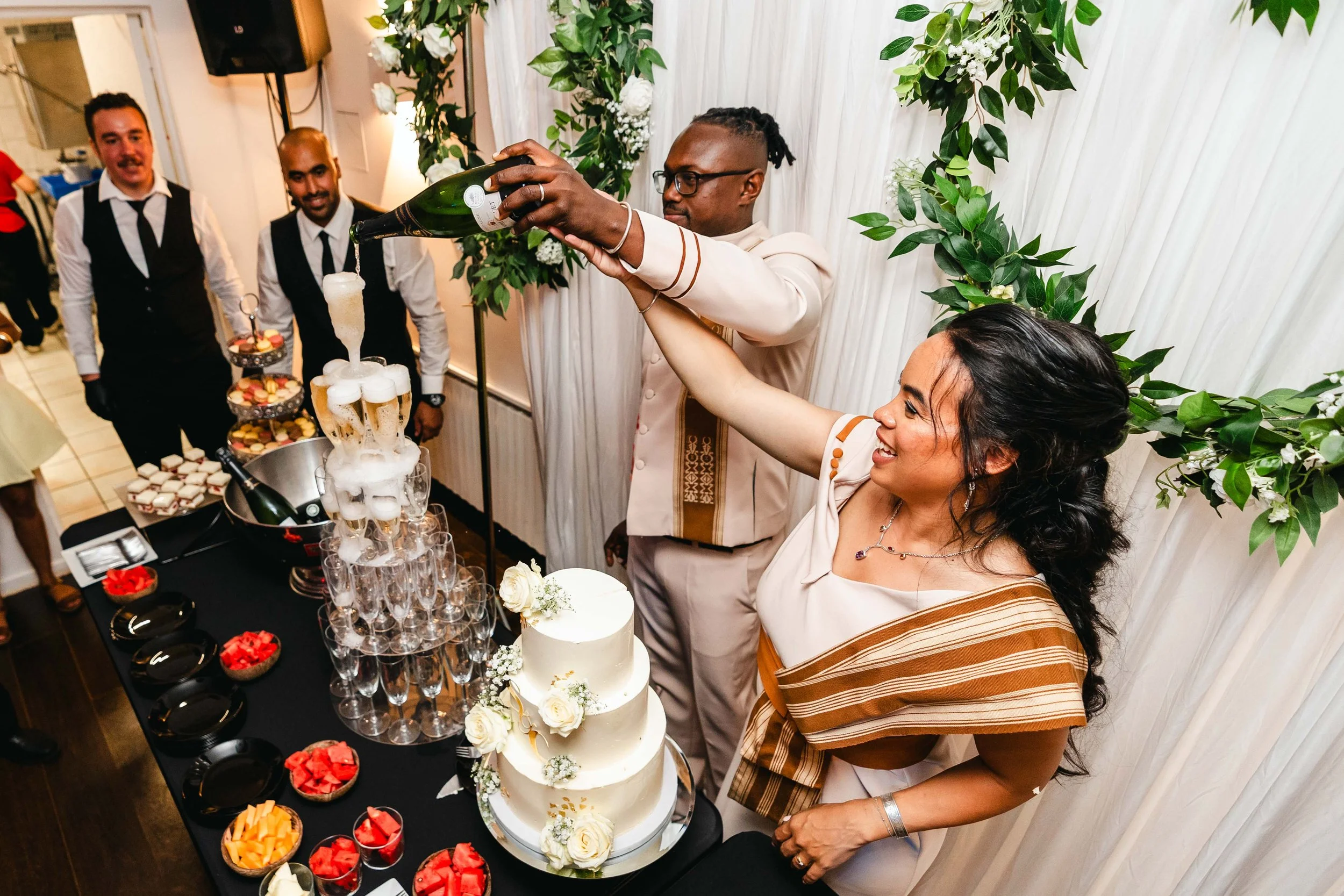 Une femme et un homme célèbrent un mariage, ils versent du champagne dans une pyramide de coupes à champagne, entourés de friends et d'une décoration florale.