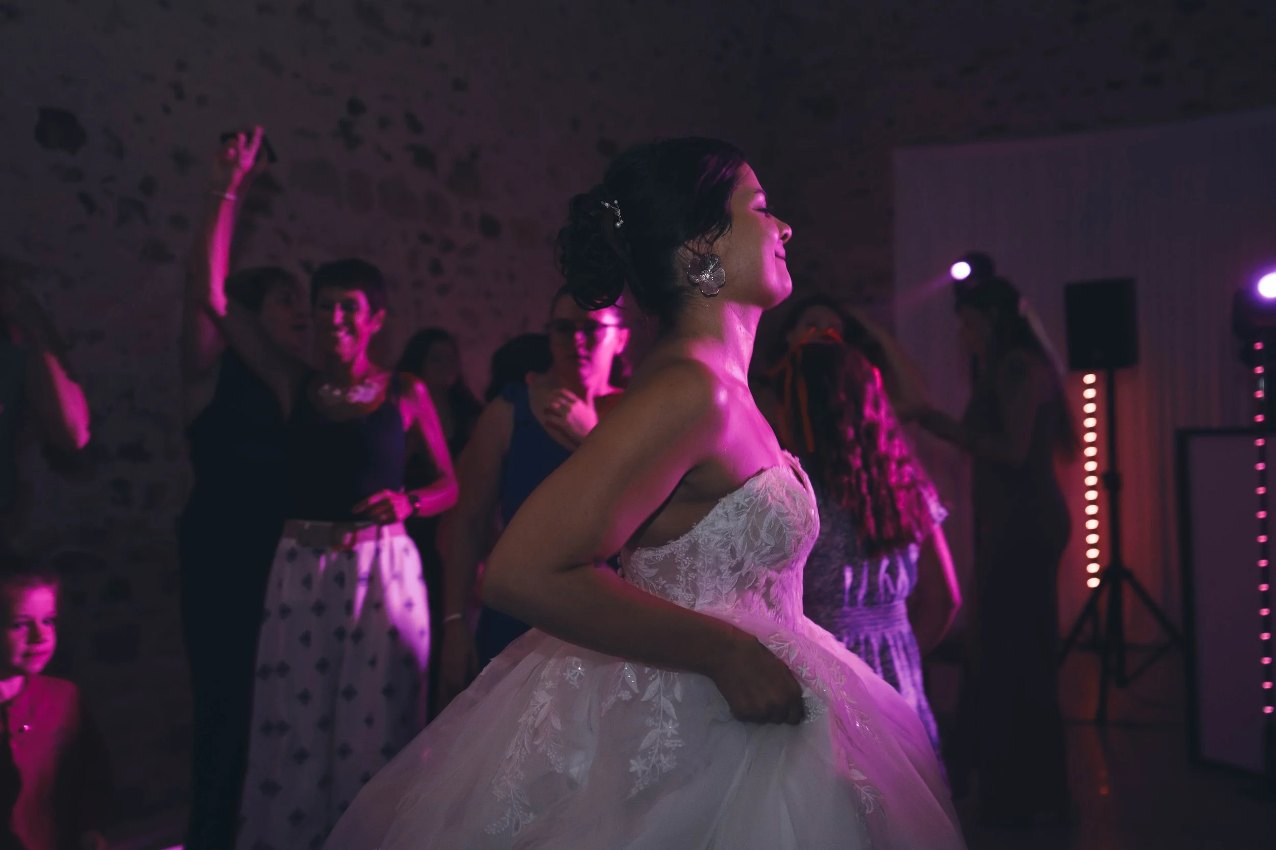 Femme en robe de mariage dans une danse lors d'une réception avec d'autres invités dans une ambiance sombre avec éclairage violet.