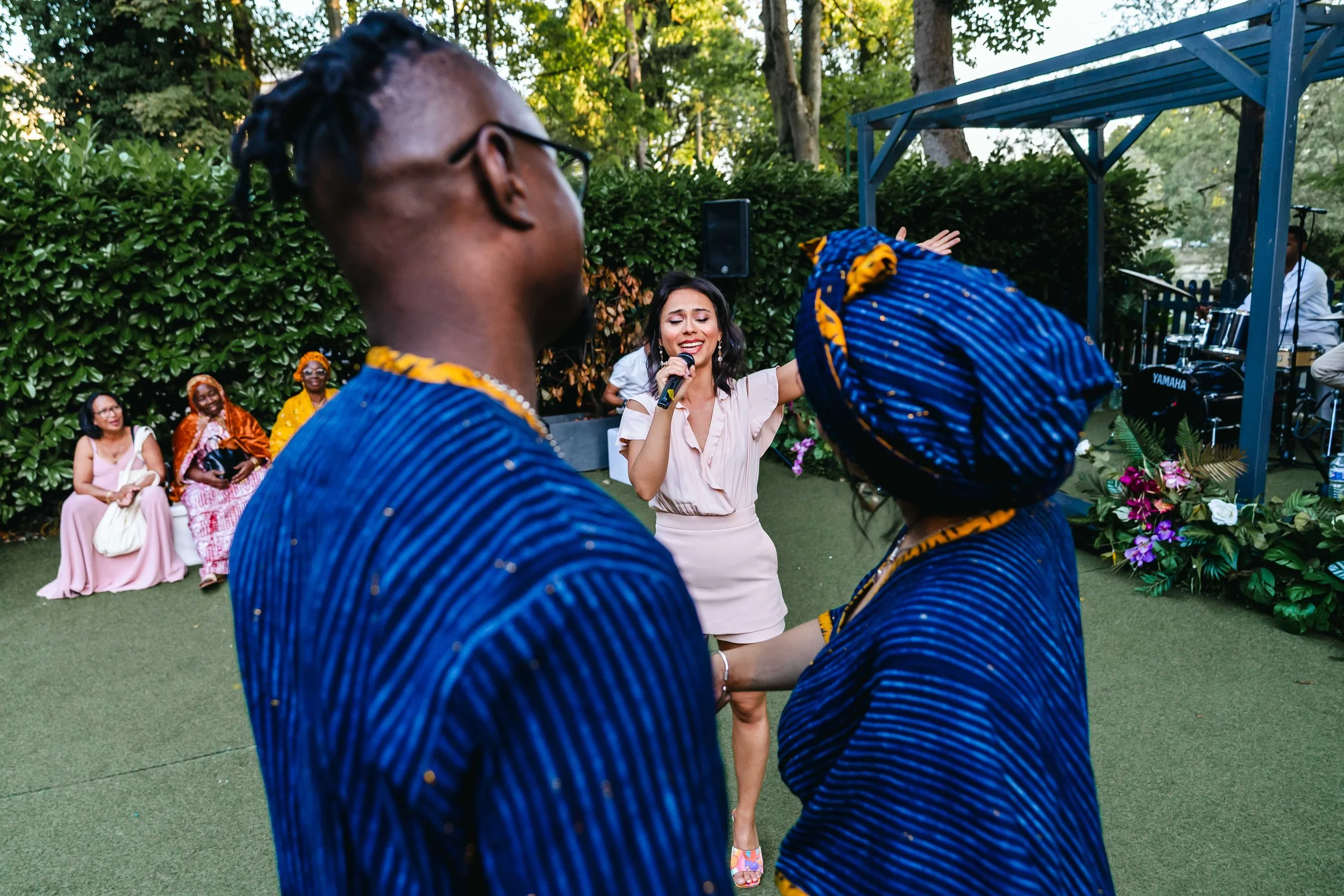 Une chanteuse en train de chanter avec un micro lors d'une célébration extérieure, entourée de plusieurs personnes, dont deux en vêtements traditionnels africains. Un groupe de musiciens joue sur scène en arrière-plan.
