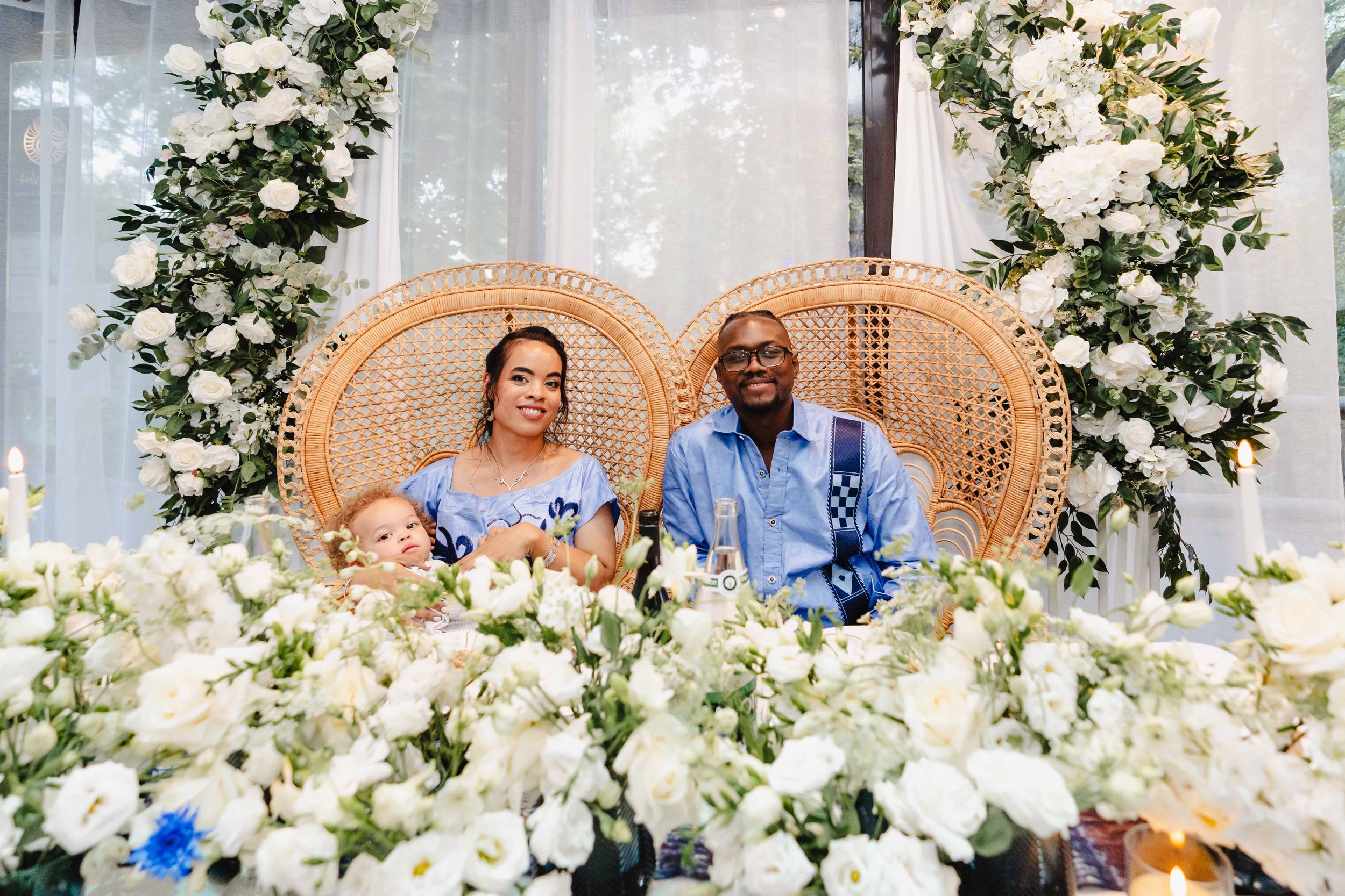 Un groupe de trois personnes, deux adultes et un enfant, assis à une table décorée de fleurs blanches, avec un arrière-plan de fleurs et de draperies blanches dans une ambiance lumineuse.