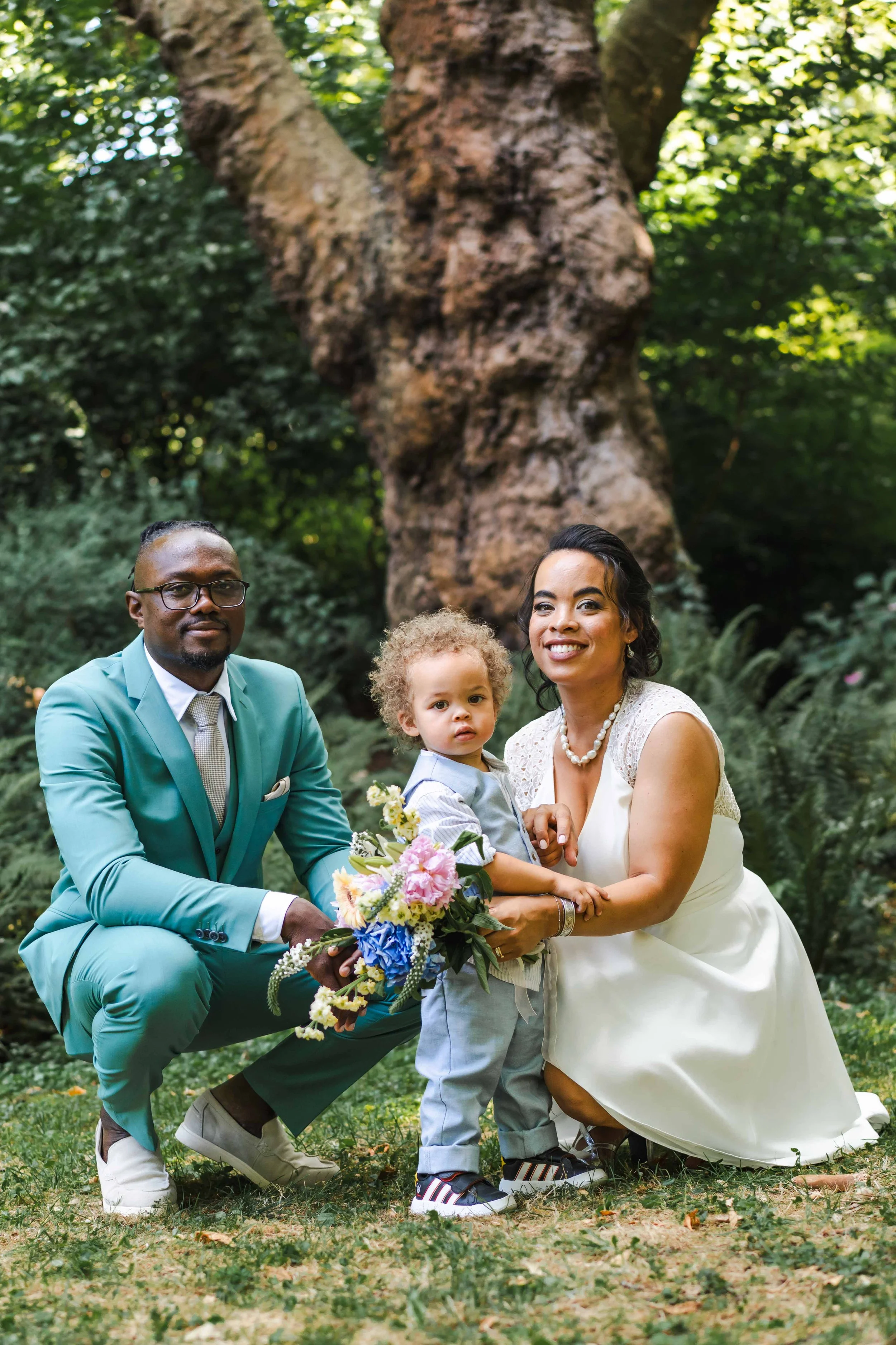 Un couple avec un enfant posant pour une photo dans un parc en forêt, lors d'une cérémonie ou d'un mariage, avec un arbre en arrière-plan.