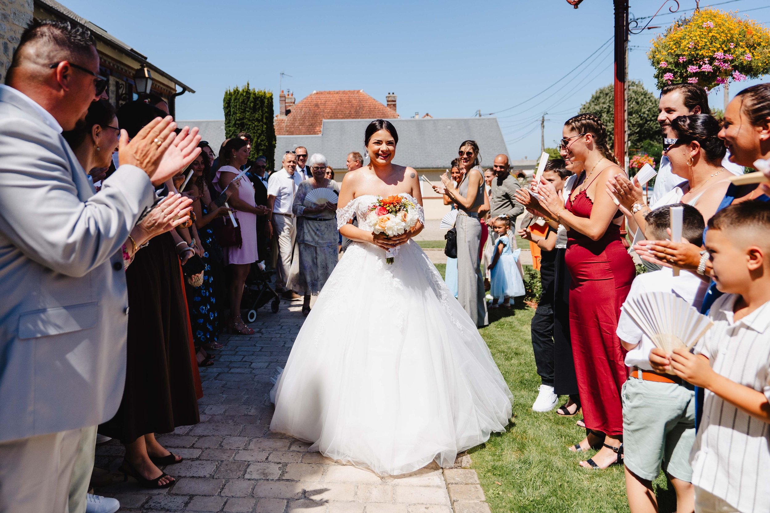 Une mariée en robe blanche souriante, tenant un bouquet de fleurs, marche dans une rue pavée entourée d'amis et de famille qui la félicitent et l'applaudissent en plein jour ensoleillé devant la mairie de Bagneux Sur Loing.