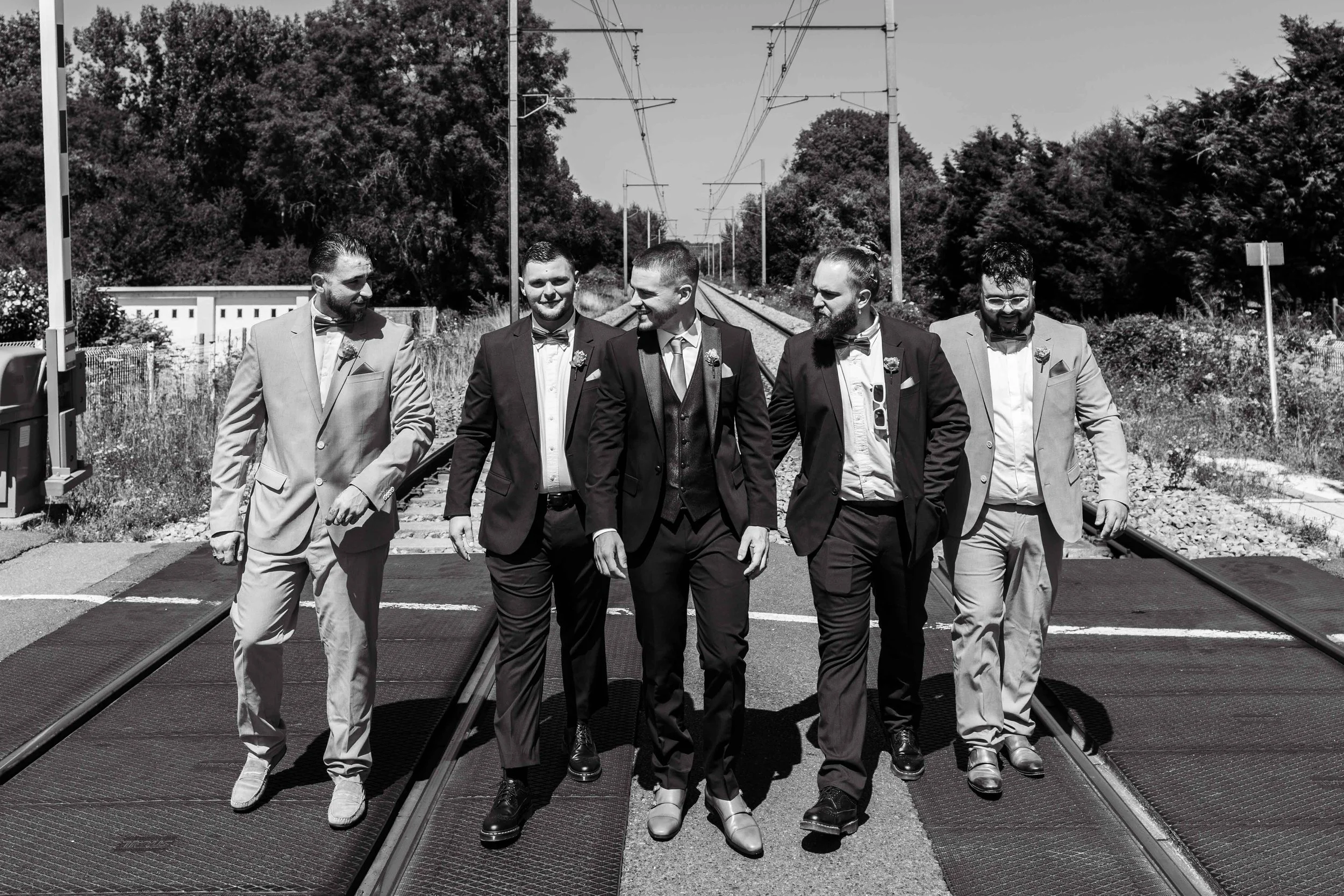 Un groupe de six hommes habillés en costumes élégants, marchant sur une voie ferrée en plein jour. L'image est en noir et blanc.