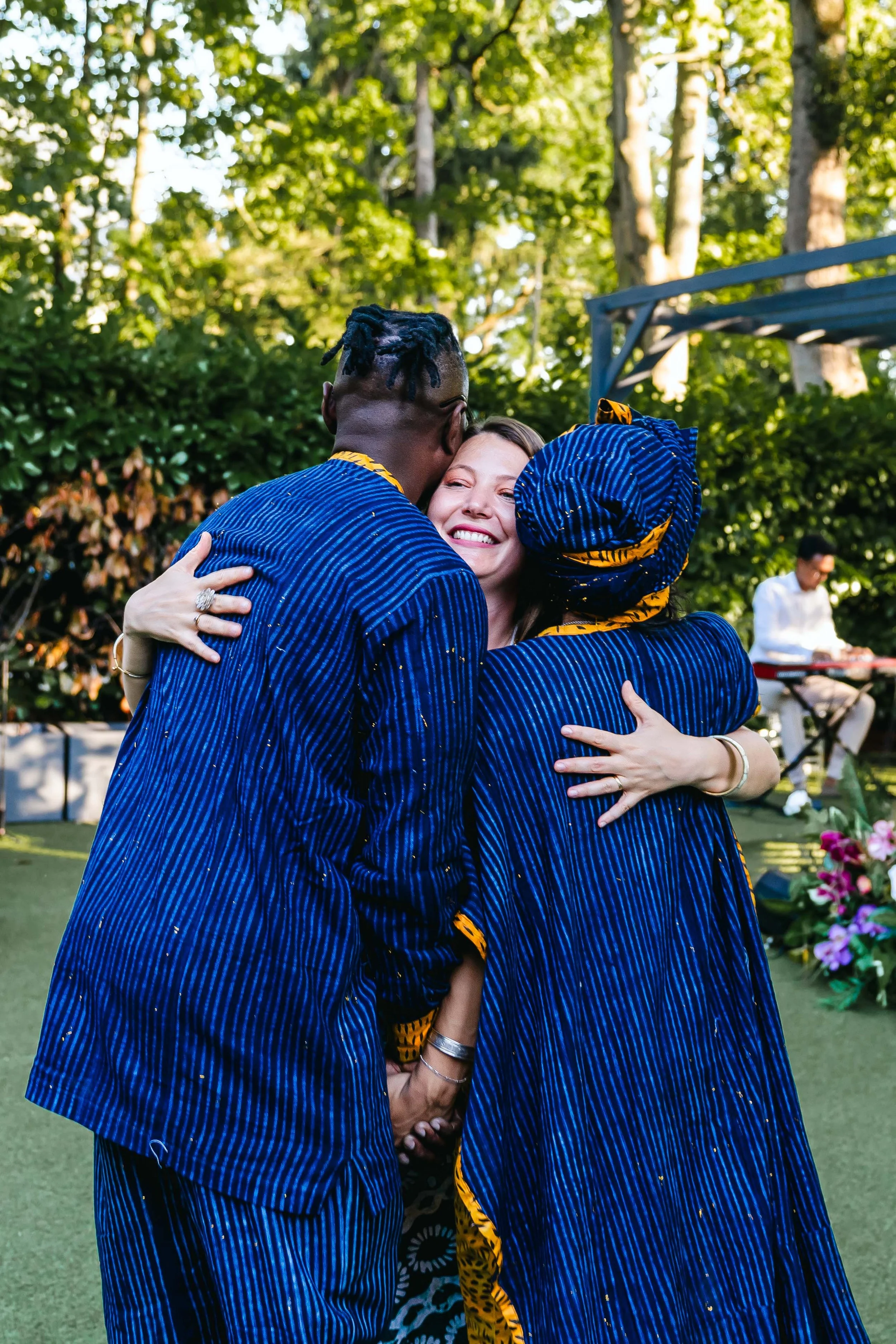 Trois personnes s'embrassent et se sourient dans un jardin, avec un musicien jouant du clavier en arrière-plan.