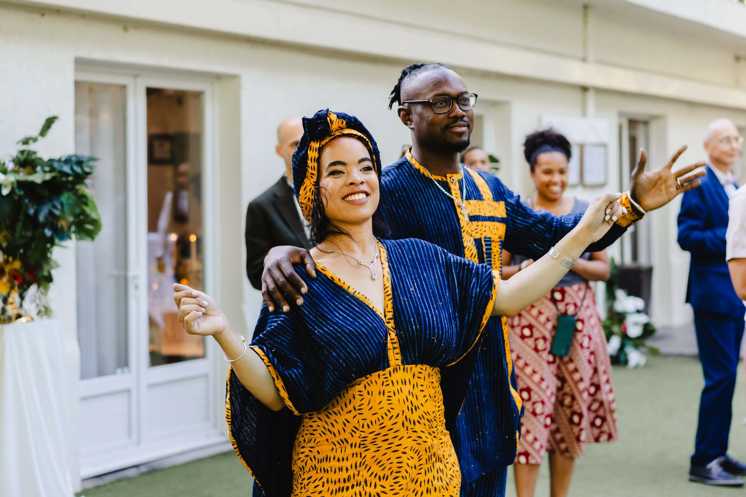 Un groupe de personnes en vêtements traditionnels africains, dans une célébration ou un rassemblement. Deux personnes au premier plan sourient, la femme porte une tenue en tissu bleu et orange, et l'homme porte une tenue coordonnée. D'autres personne