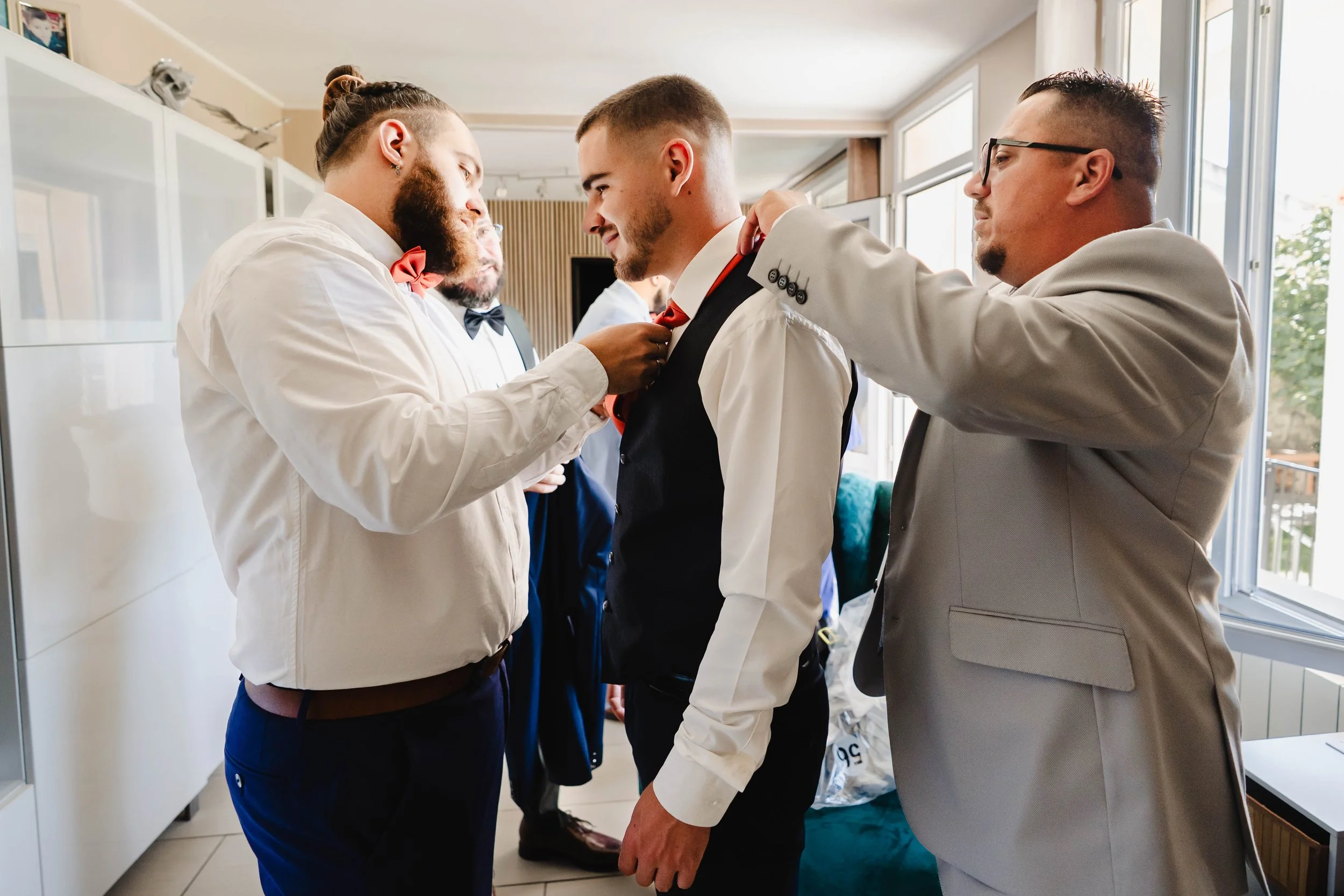 Un groupe d'hommes habillés en costumes se prépare, avec deux hommes aidant un à attacher sa cravate rouge, dans une pièce bien éclairée par de grandes fenêtres.