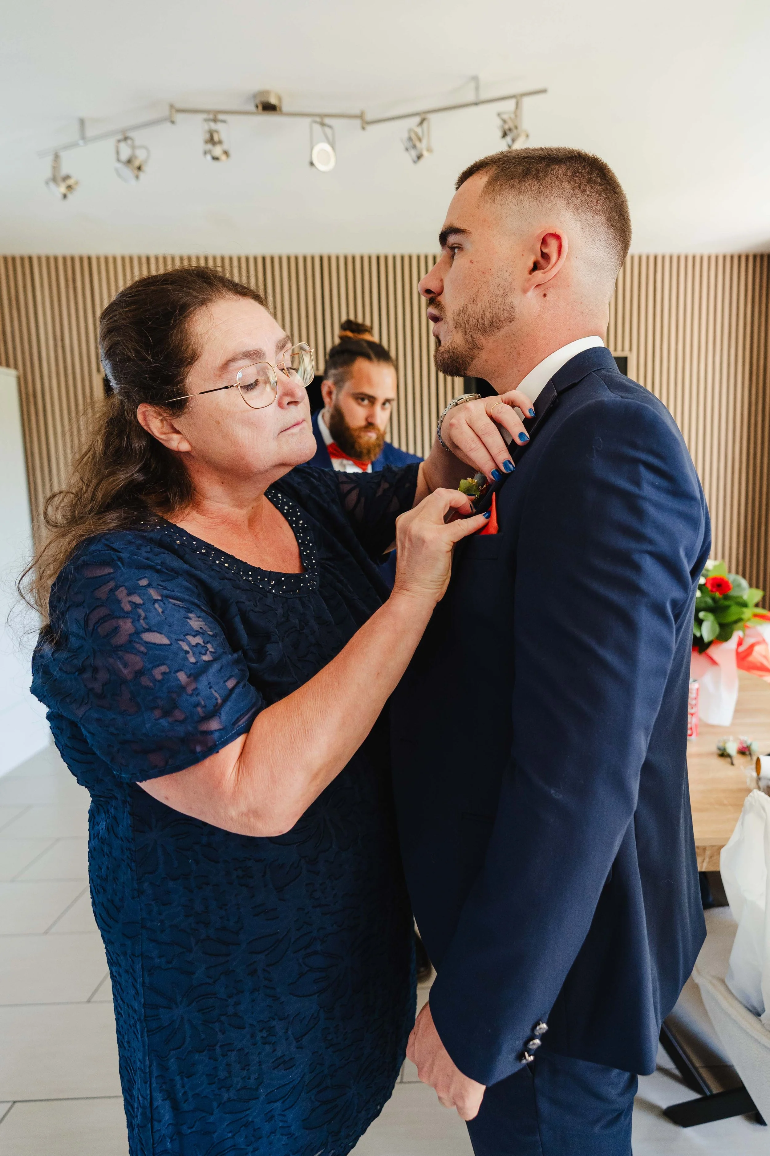 Une femme ajuste la boutonnière d'un jeune homme habillé en costume bleu, probablement lors d'une cérémonie ou d'un mariage.