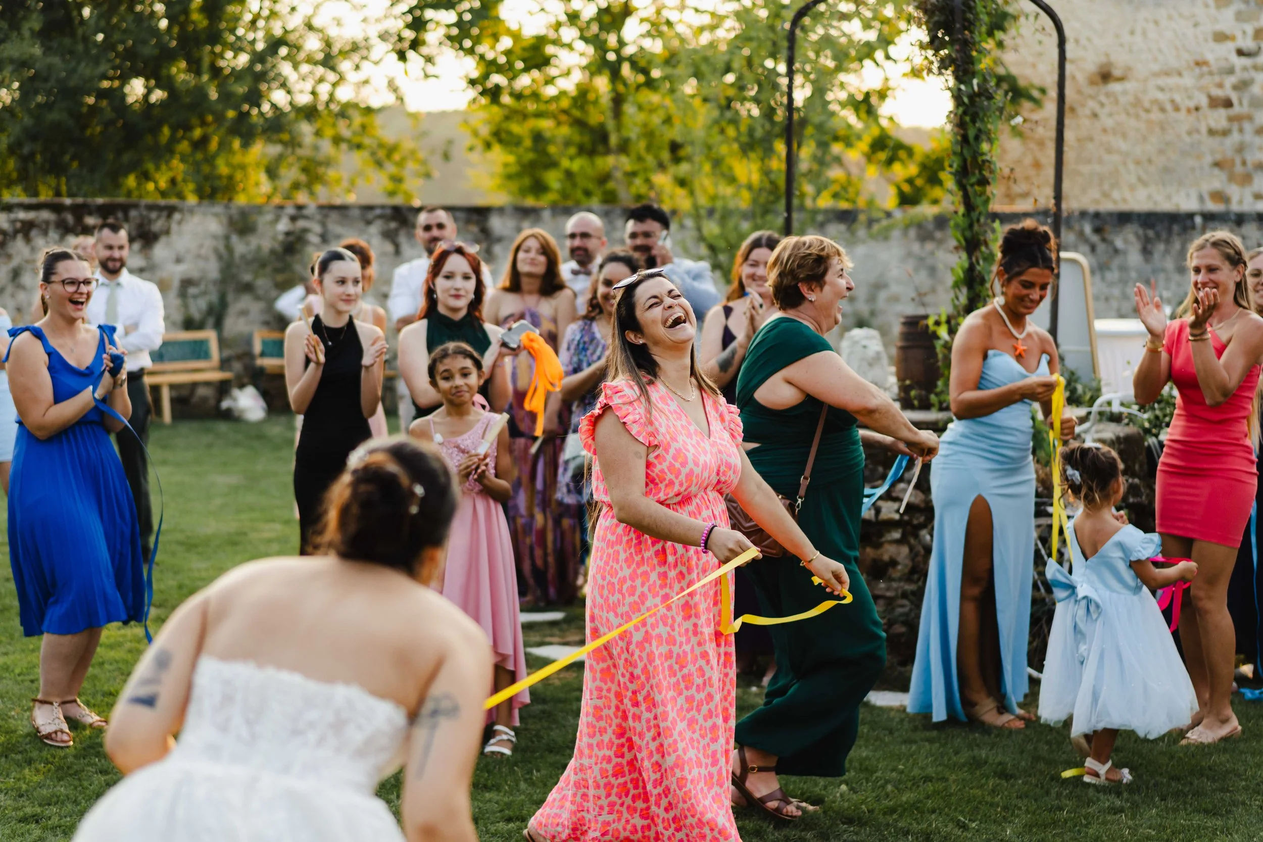 Groupe de personnes souriantes et riant lors d'une fête en plein air, avec des femmes en robes colorées et des enfants, dans un jardin en soirée.