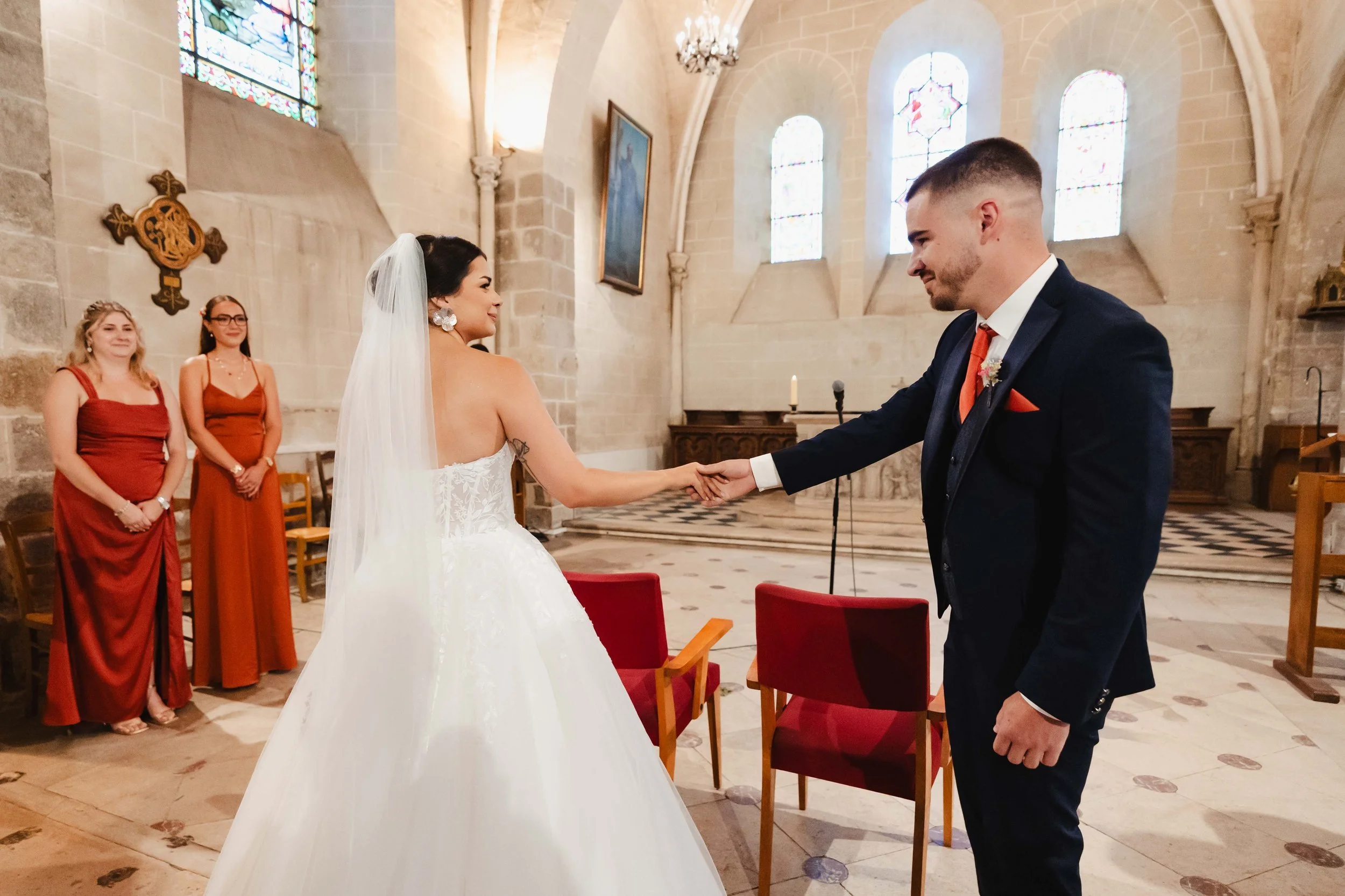 Un mariage dans une église, le couple échangeant leurs vœux, avec deux femmes en robe rouge en arrière-plan.