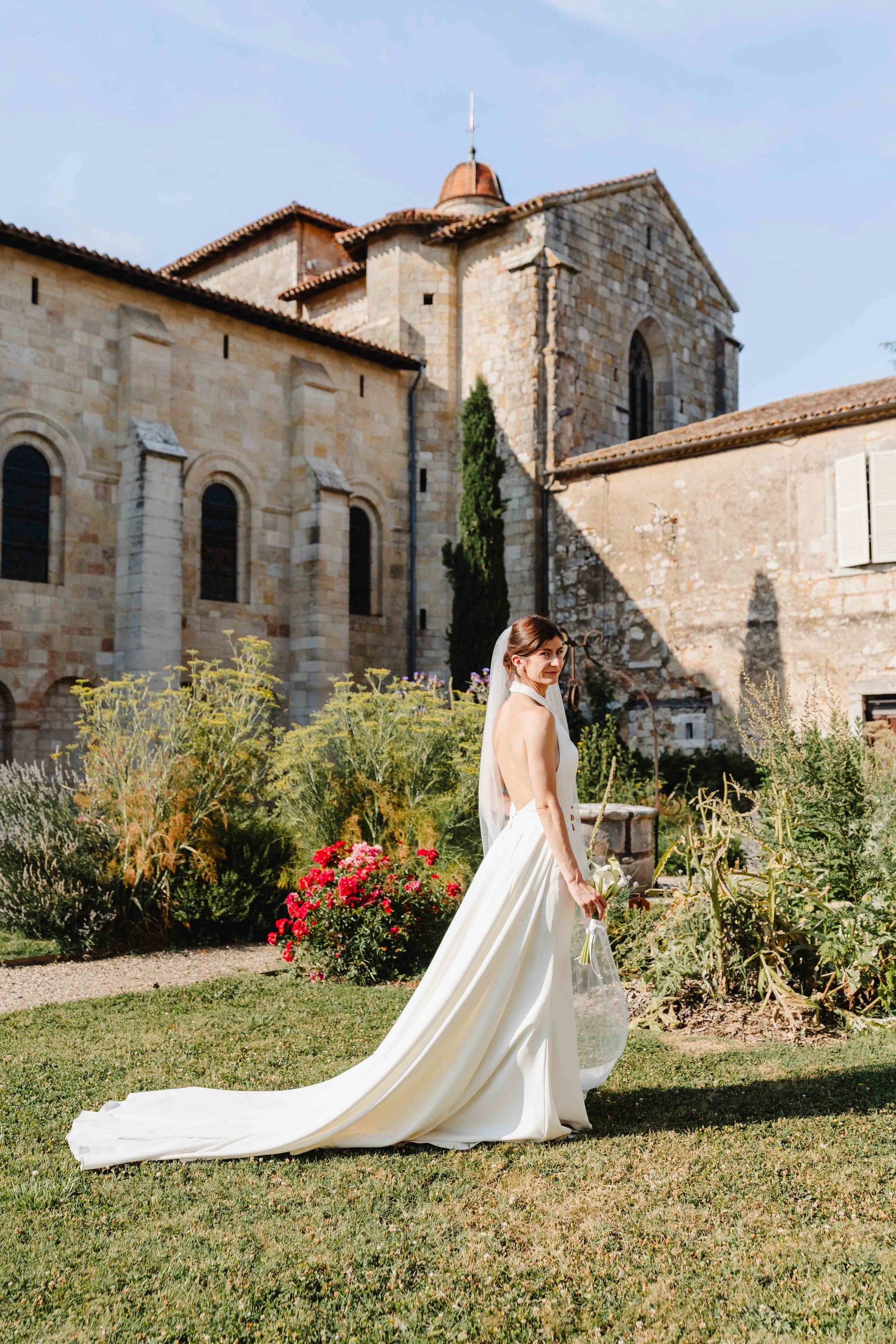 Une mariée en robe blanche avec traîne, tenant un bouquet, dans un jardin, devant l'église de Moirax