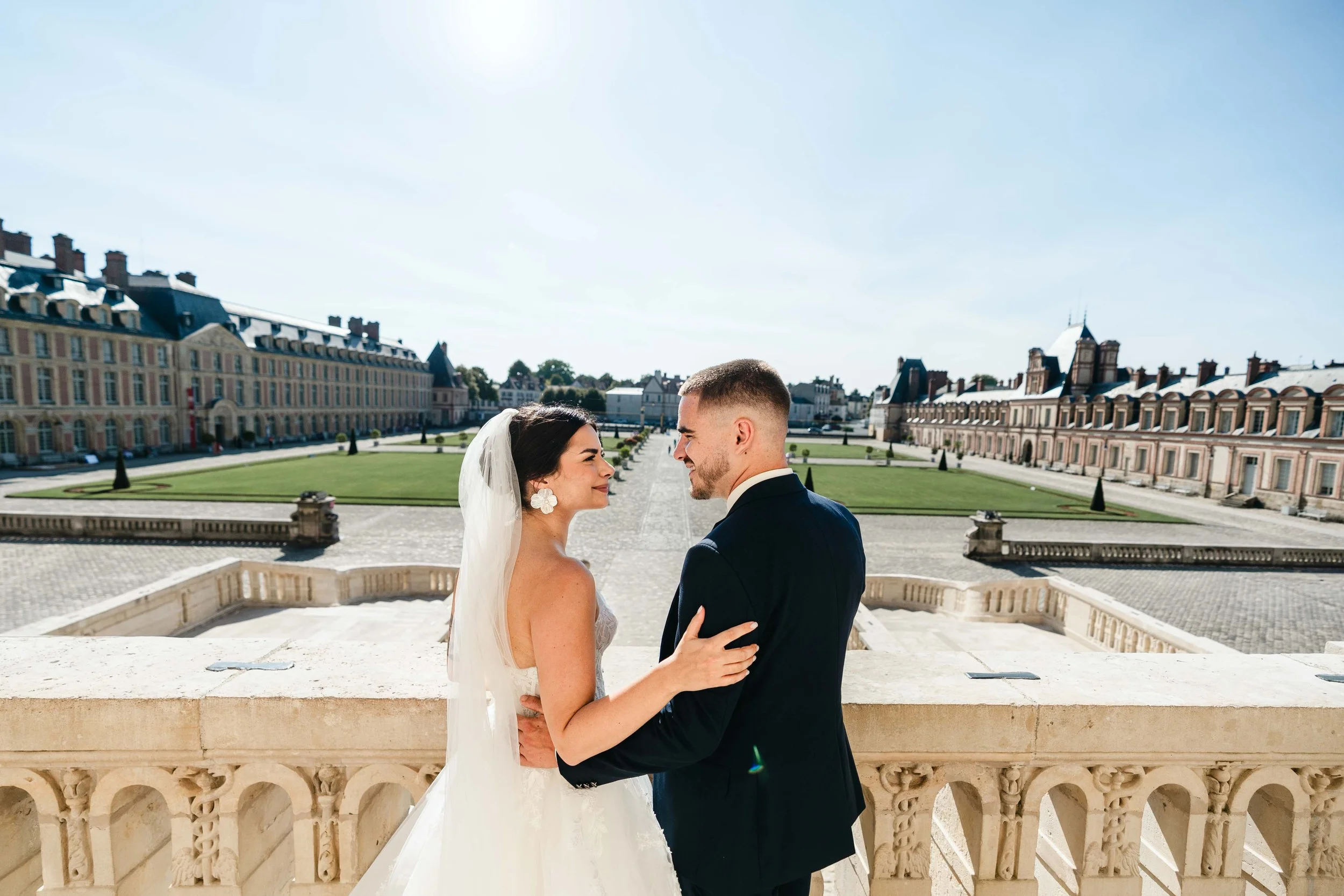 Un couple de mariés souriants, la femme en robe de mariage et le homme en costume, se tenant la main sur le balcon en pierre du château de Fontainebleau, avec le jardin en arrière-plan sous un ciel clair.